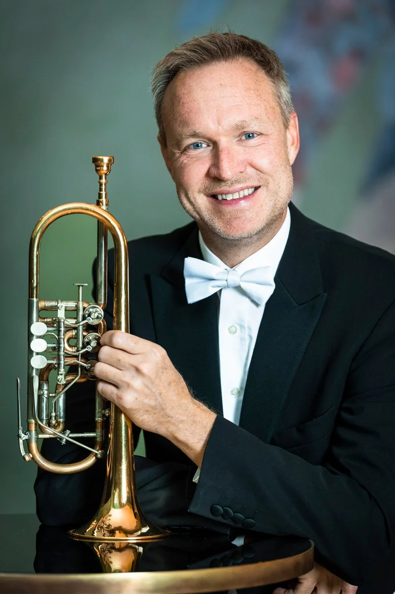 A man in a tuxedo holding a trumpet, smiling at the camera.