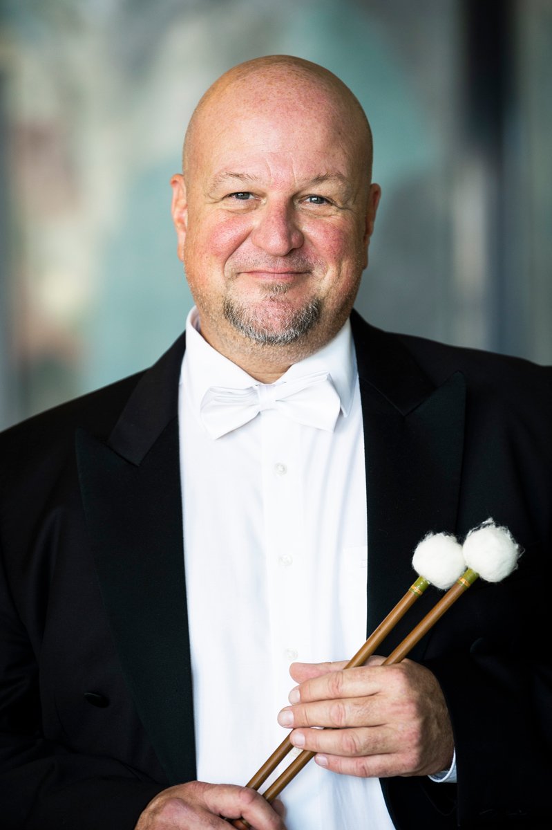 A man with a shaved head and a goatee wearing a formal tuxedo with a black jacket and a white bow tie, holding a pair of drumsticks with white cotton tips, standing against a blurred background.