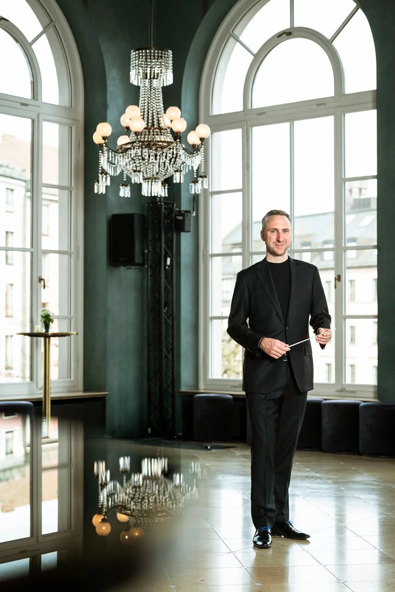 A man in a black suit standing in a brightly lit room with large arched windows, a chandelier, and a reflection on the floor.