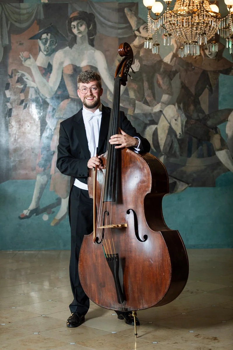 A young man in a black tuxedo holding a large double bass in a room with a mural of dancers and a chandelier hanging from the ceiling.