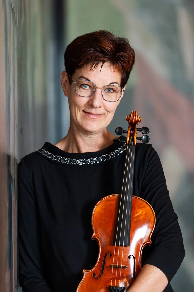 A woman with short dark hair and glasses holding a violin and smiling
