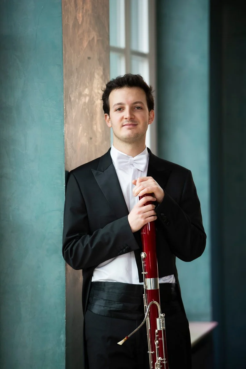 A young man in formal attire holding a bassoon, standing indoors near a window with green and brown textured walls.