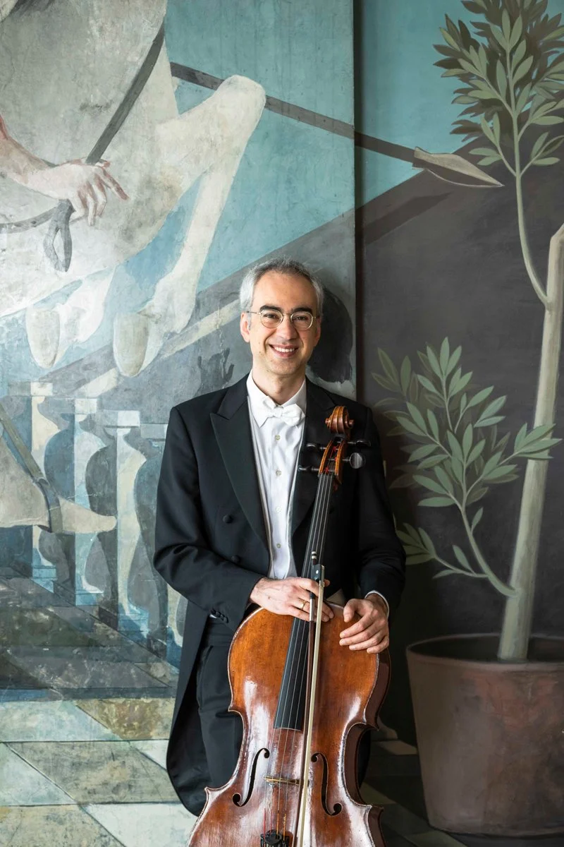 A man in a tuxedo holding a cello, smiling and standing in front of a mural with a painting of a person climbing stairs and a potted plant.
