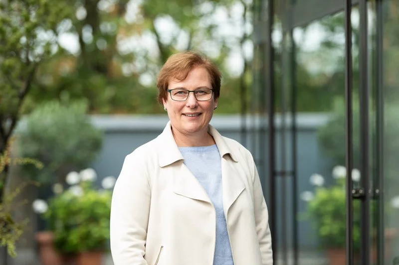 A woman with short light brown hair, glasses, and a beige coat standing outdoors in front of a glass building with greenery in the background.