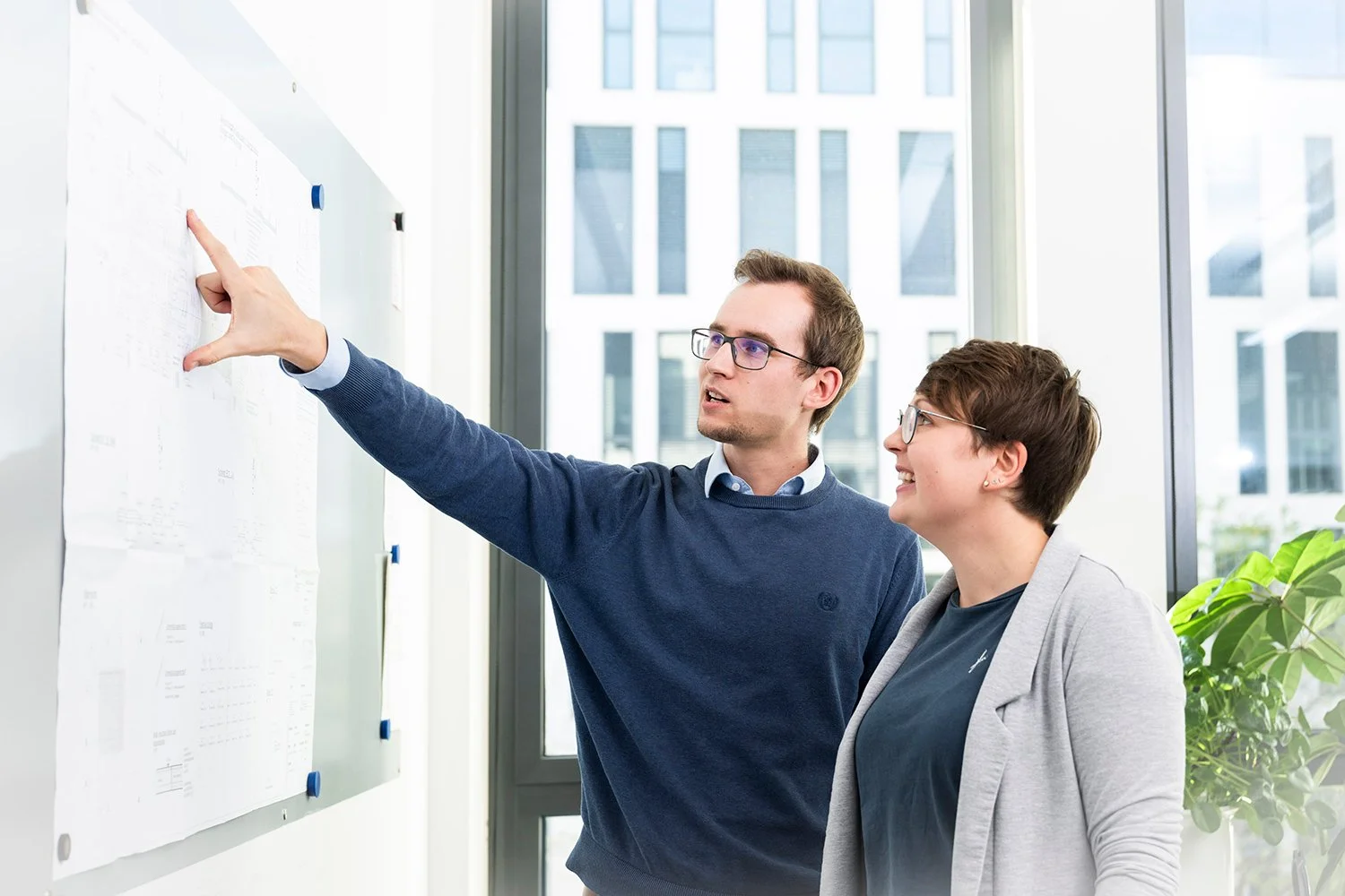 Two people, a man and a woman, standing in front of a whiteboard with papers, the man pointing at it while explaining content, in a bright office with large windows.