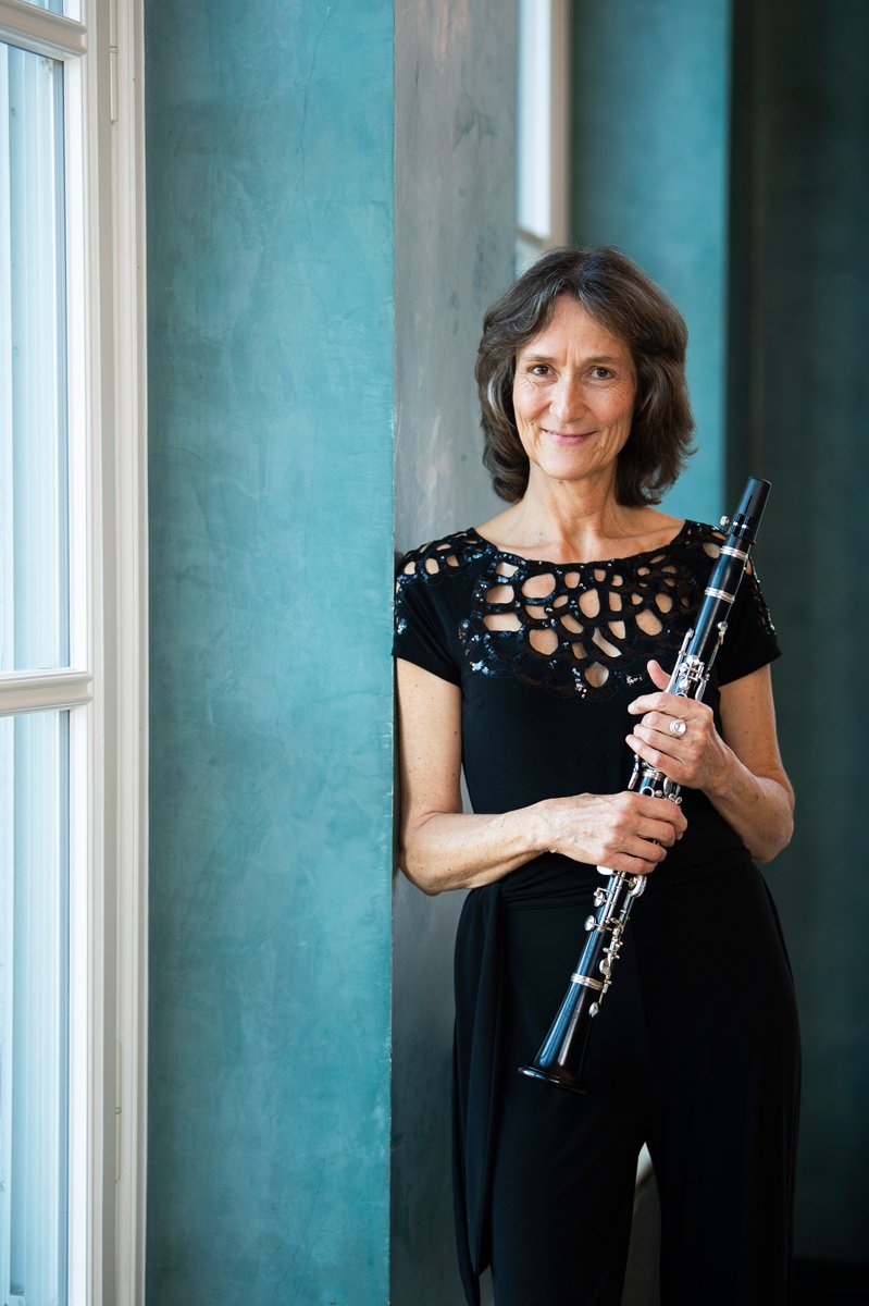 A woman standing indoors near a window, holding a clarinet, and smiling at the camera.