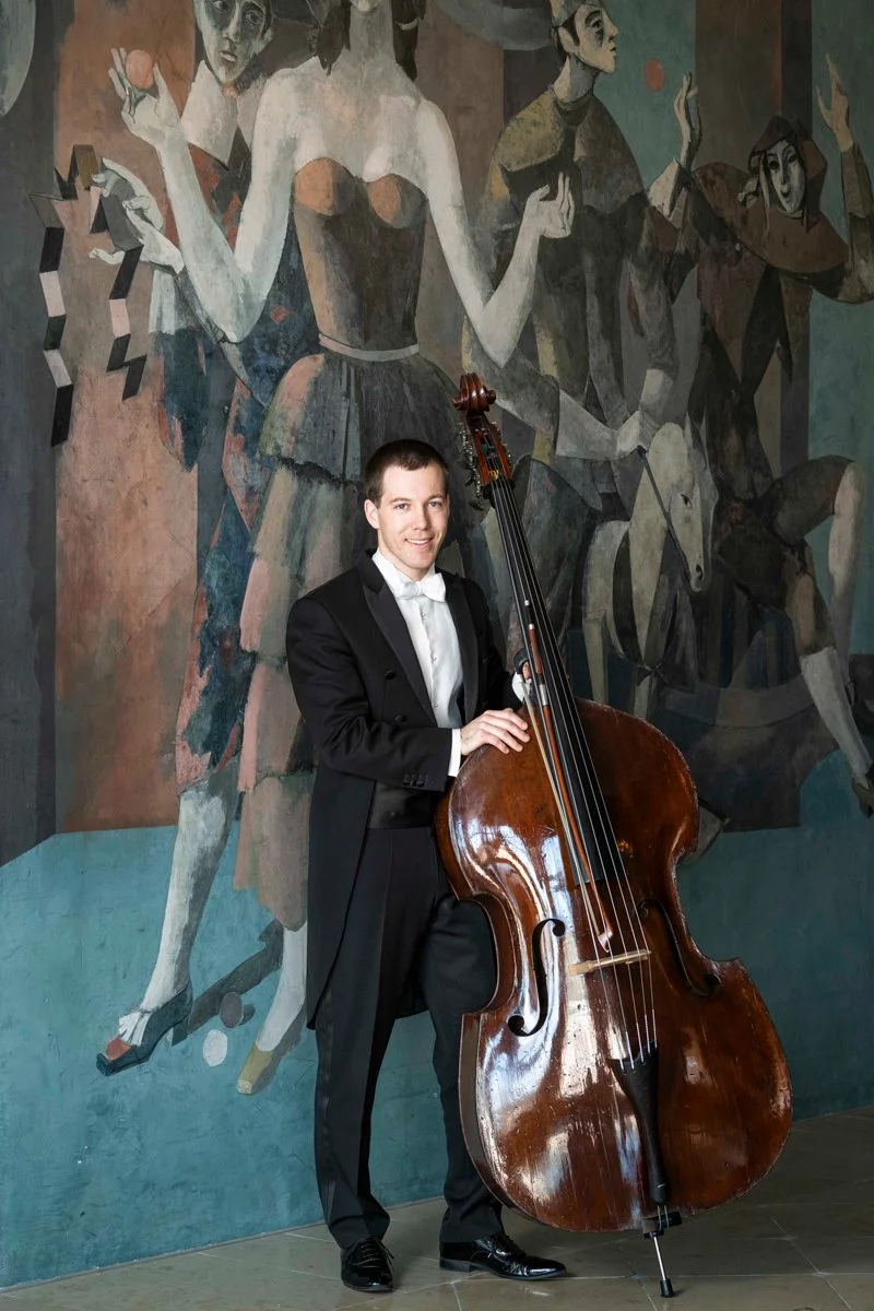 A man in a black tuxedo with a white bow tie standing beside a double bass in front of a large abstract mural painting.