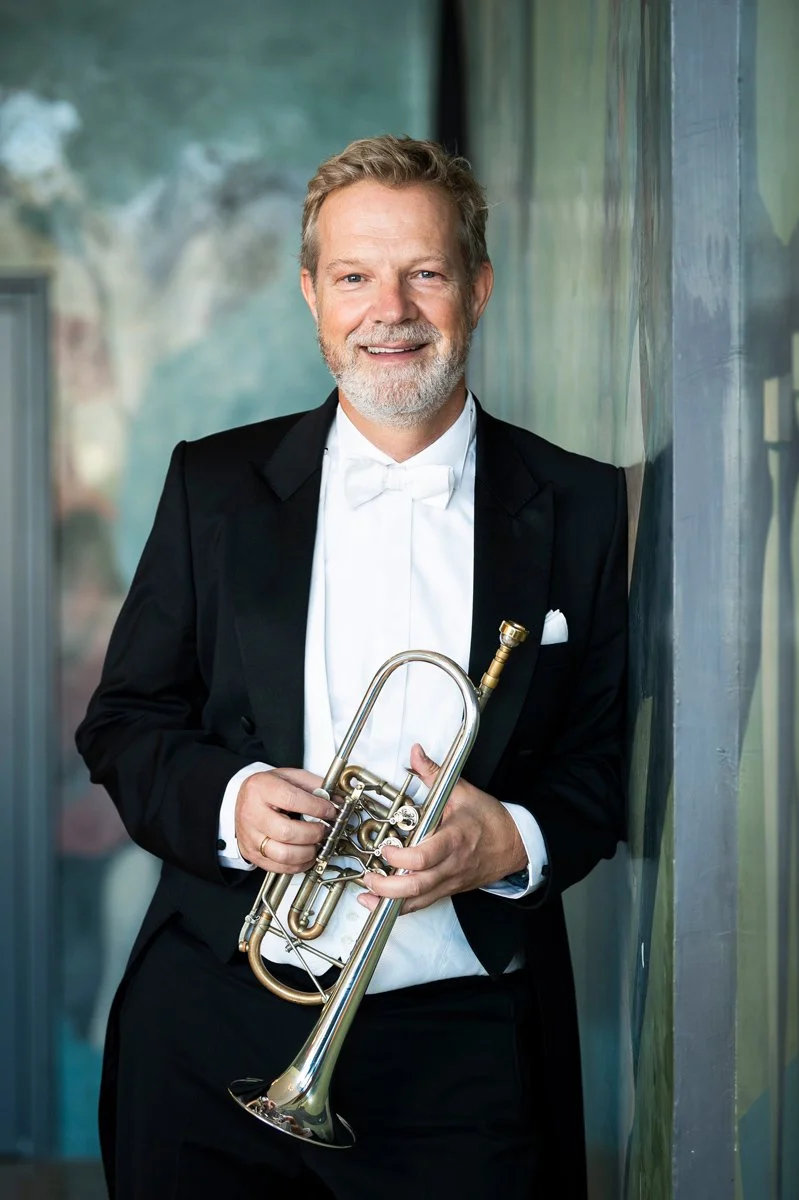 A smiling man with gray hair and beard, dressed in a black tuxedo with a white bowtie, holding a trumpet, standing next to a colorful abstract wall.
