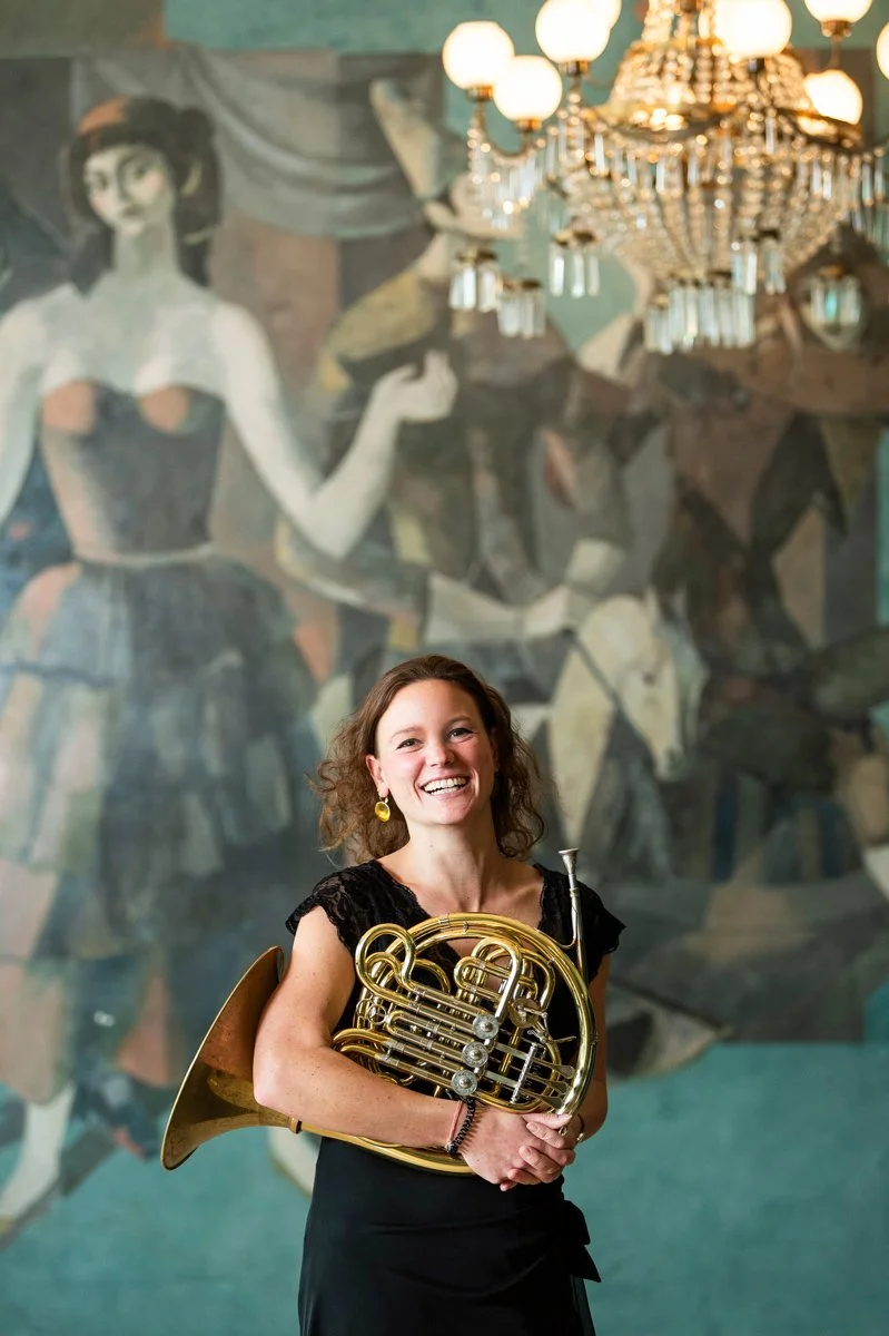 A woman with curly brown hair, wearing a black dress with lace sleeves, smiling and holding a French horn while standing in front of a painted mural of a woman in a dress and a chandelier.