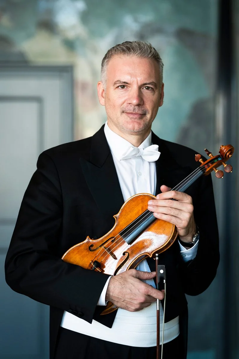 A man in formal attire, wearing a tuxedo with a white bow tie, holding a violin and a bow, standing indoors with a blurred background.