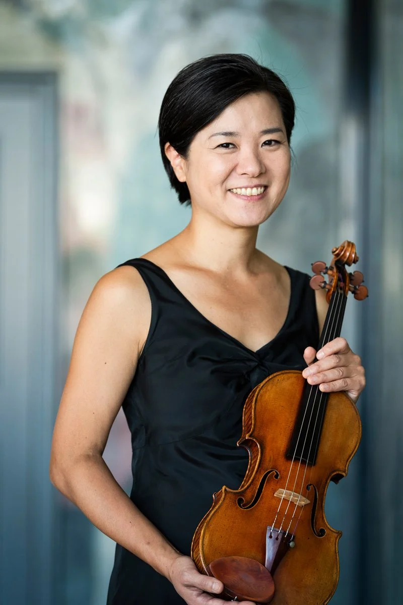 A woman with short black hair wearing a black dress holding a violin and smiling.