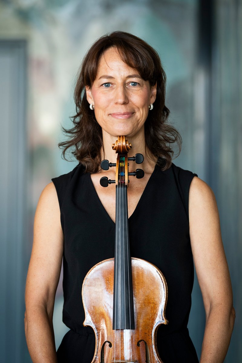 A woman with brown hair and blue eyes holding a violin in front of her chest, standing indoors with a blurred background.
