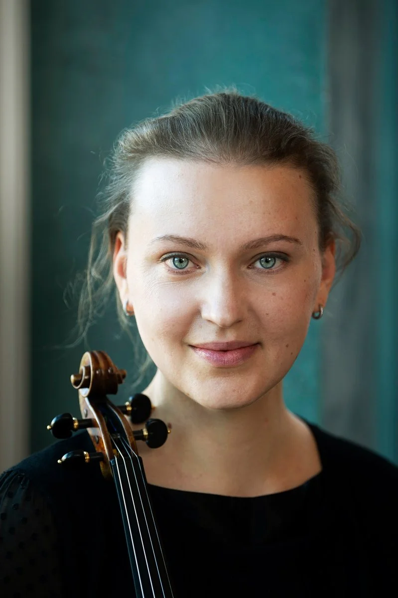 A young woman with light skin, blue eyes, and brown hair pulled back, holding a violin near her shoulder, wearing a black top, standing indoors with a blurred blue and green background.