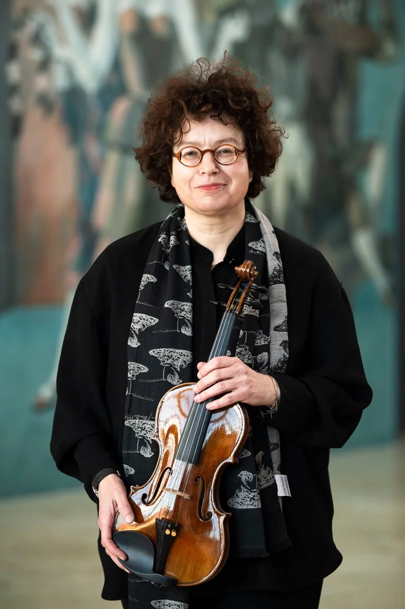 A woman with curly hair and glasses holding a violin, standing in front of an abstract background.