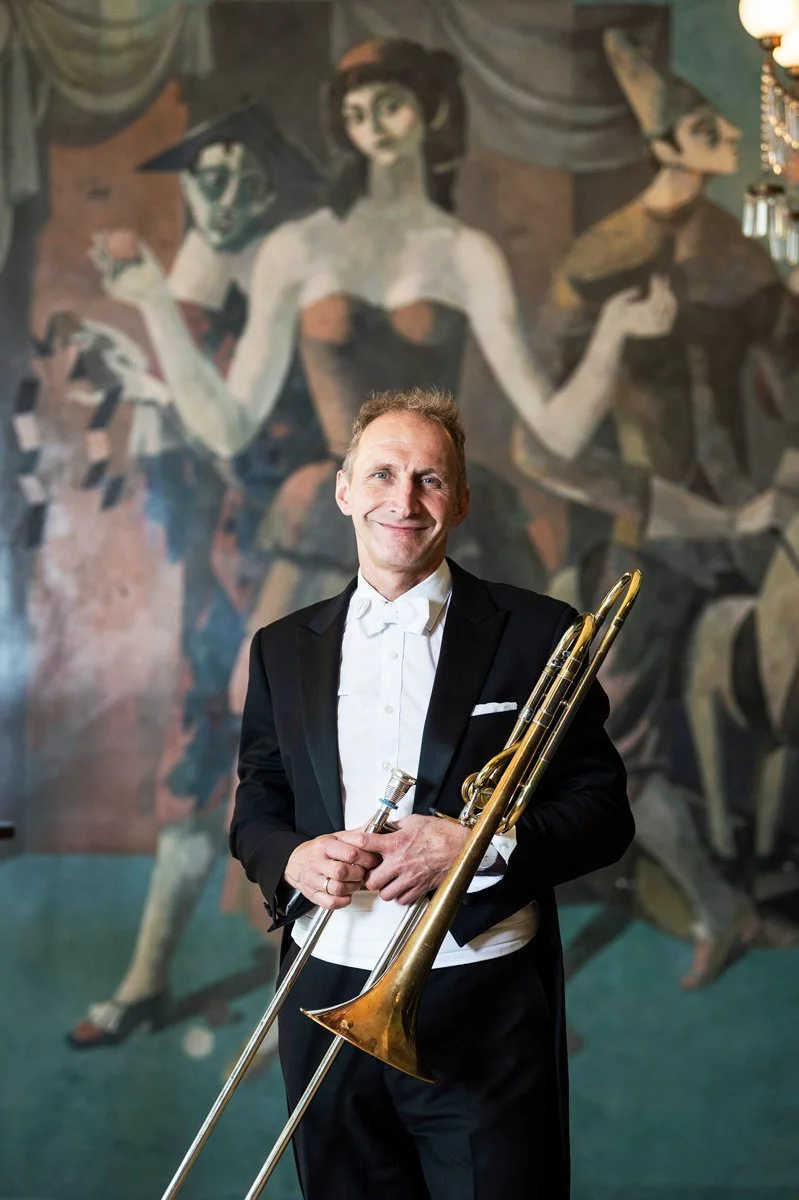 A man in a formal tuxedo holding a trombone and a small object, standing in front of a large mural featuring stylized, vintage-style women in an art deco setting.