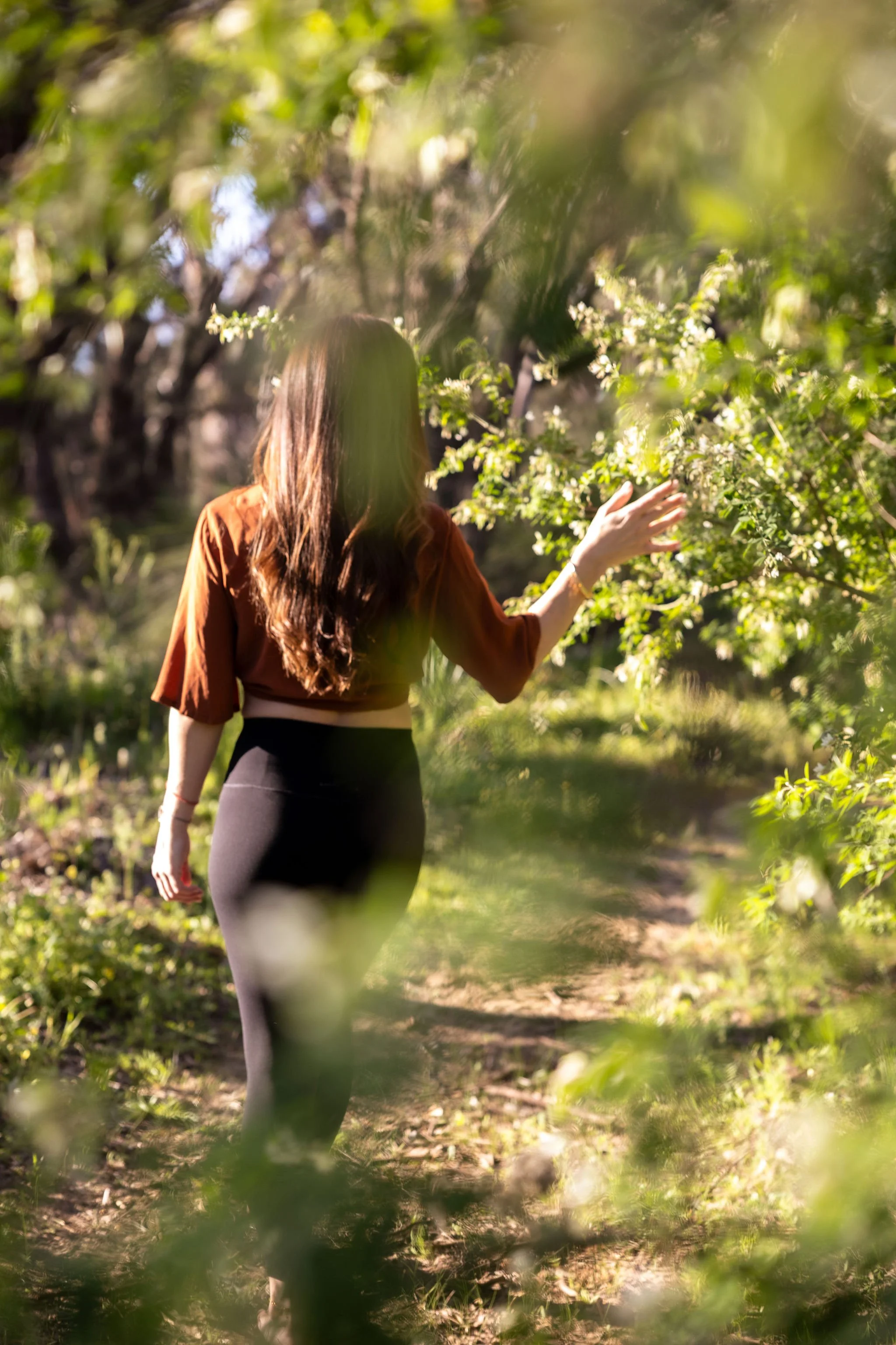 A woman with long brown hair, wearing a brown top and black leggings, walking through a lush garden or orchard with green foliage and blooming trees, sunlight filtering through the leaves.