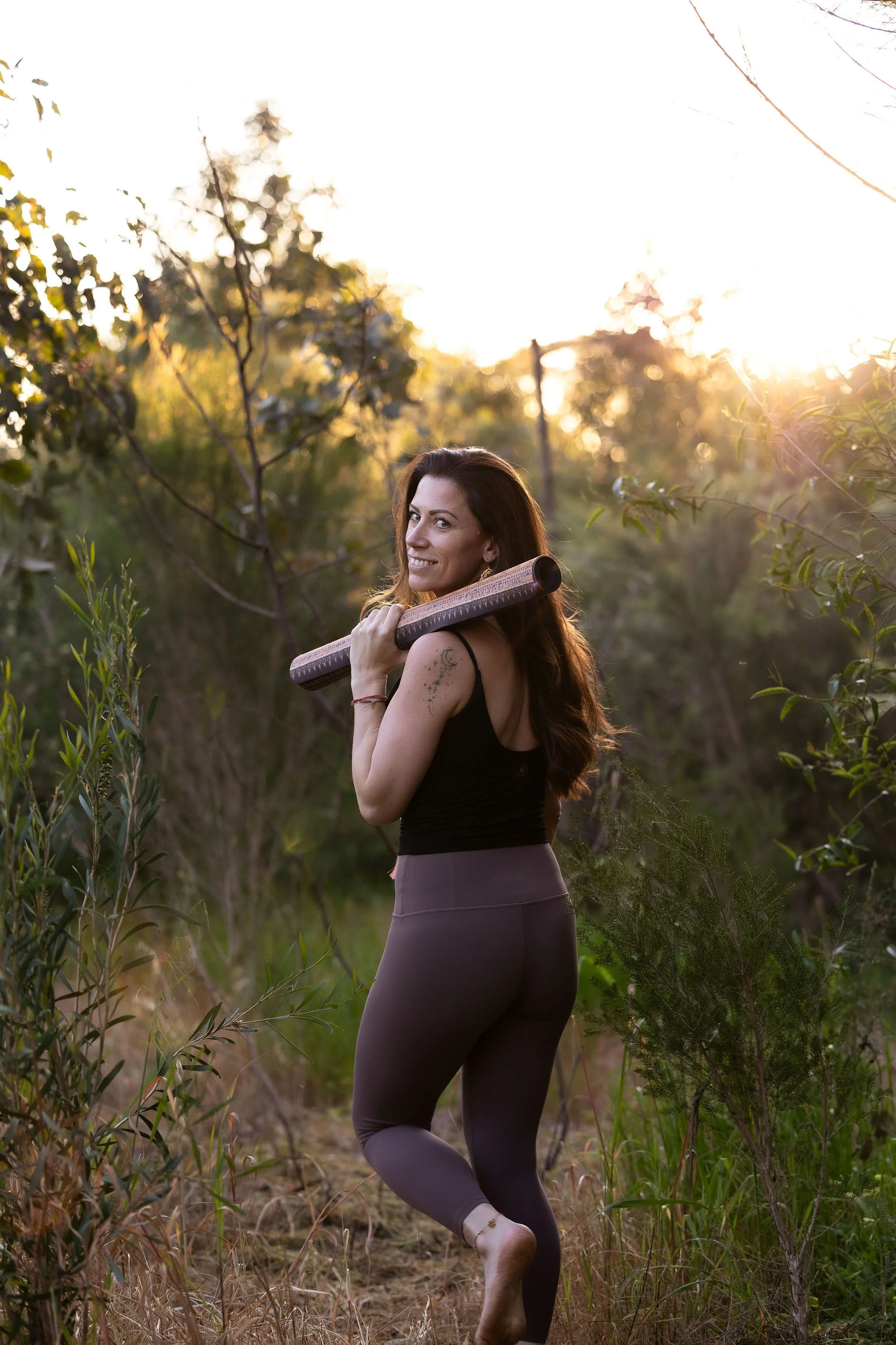 A woman with long brown hair and a tattoo on her arm, smiling and carrying a yoga mat, stands amidst green plants and trees during sunset.