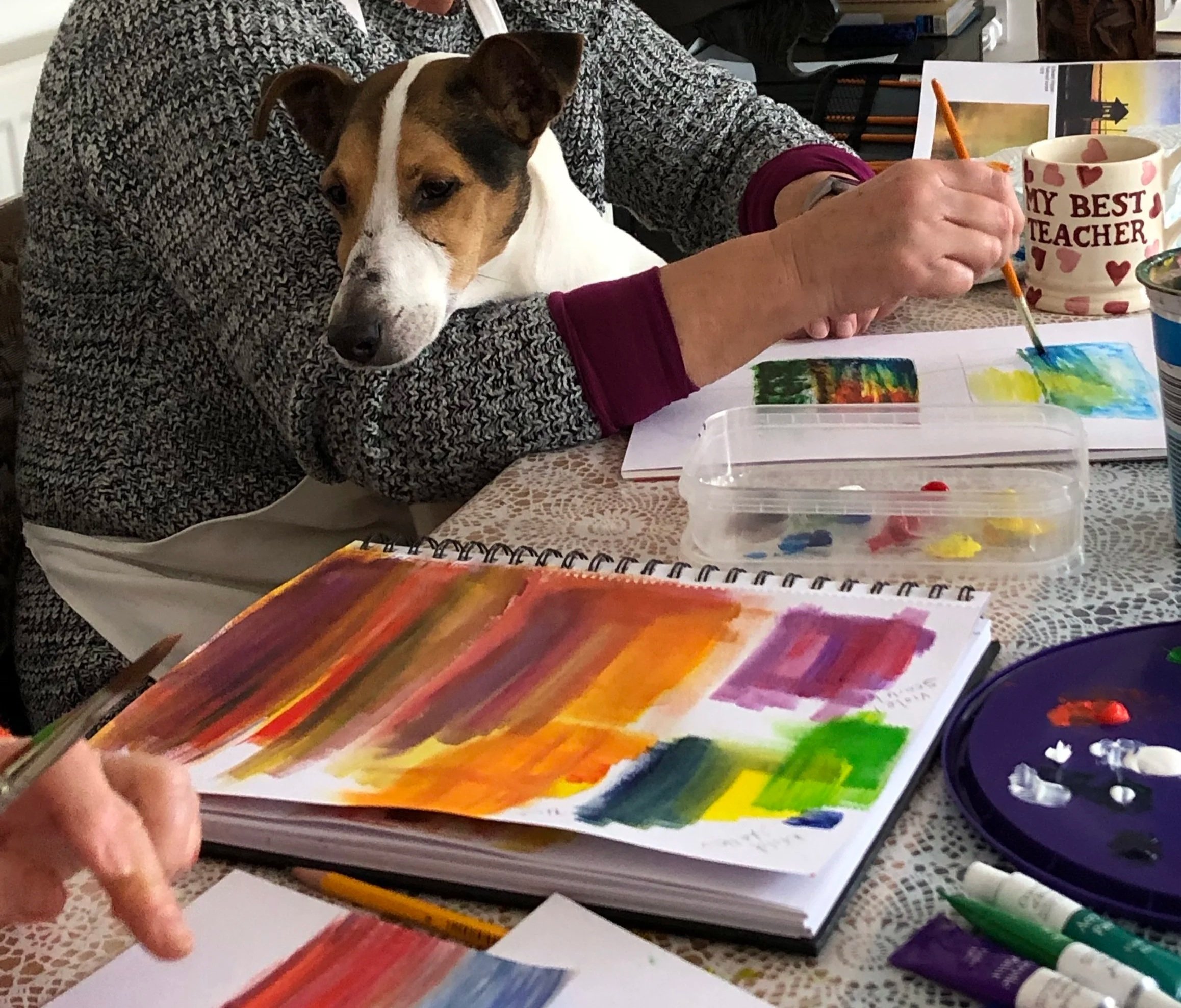 Person painting colorful abstract art with dog resting its head on their arm at a table with paints and brushes.