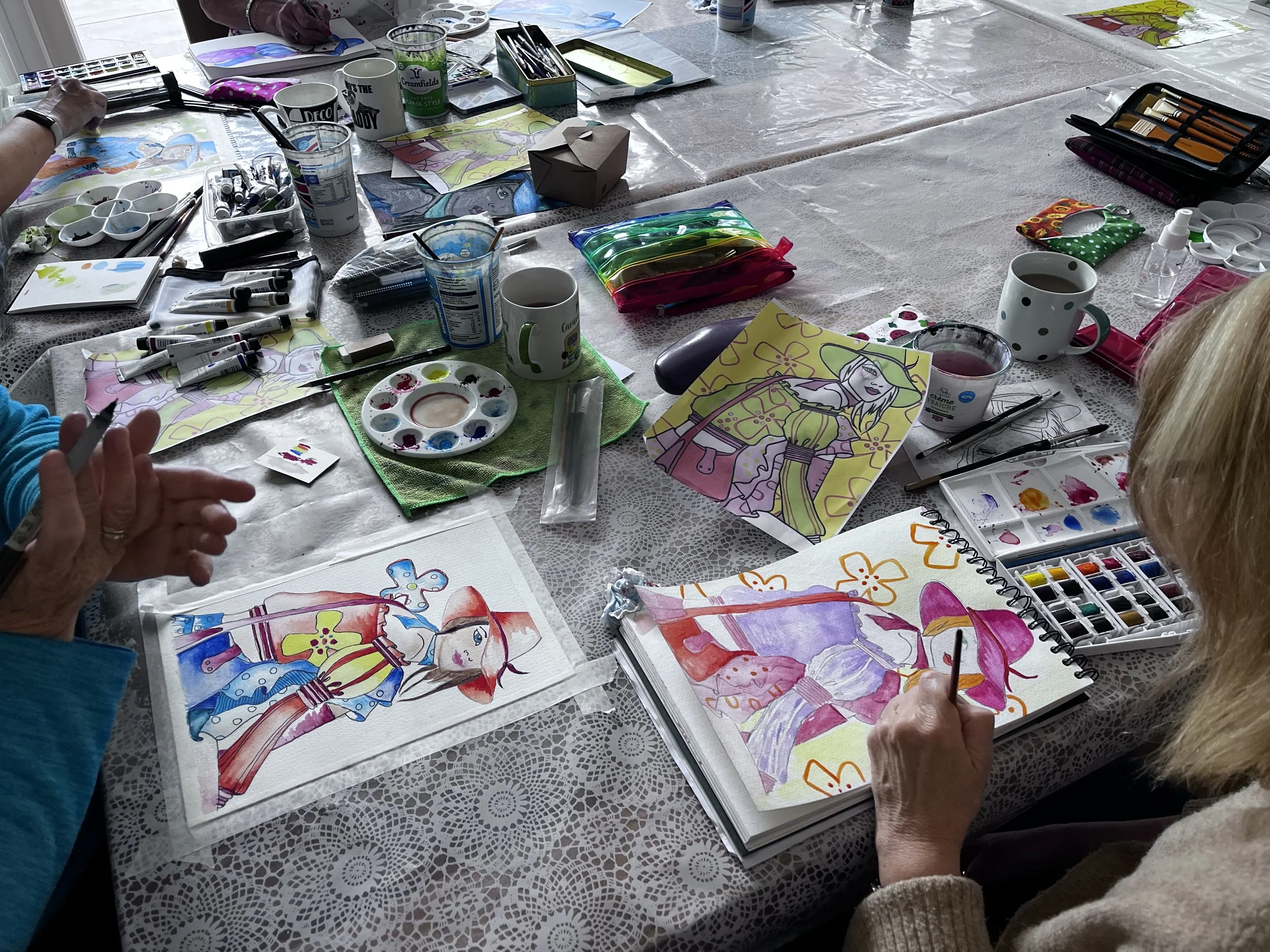 People painting colorful illustrations of women and flowers on paper at a table with art supplies and cups.