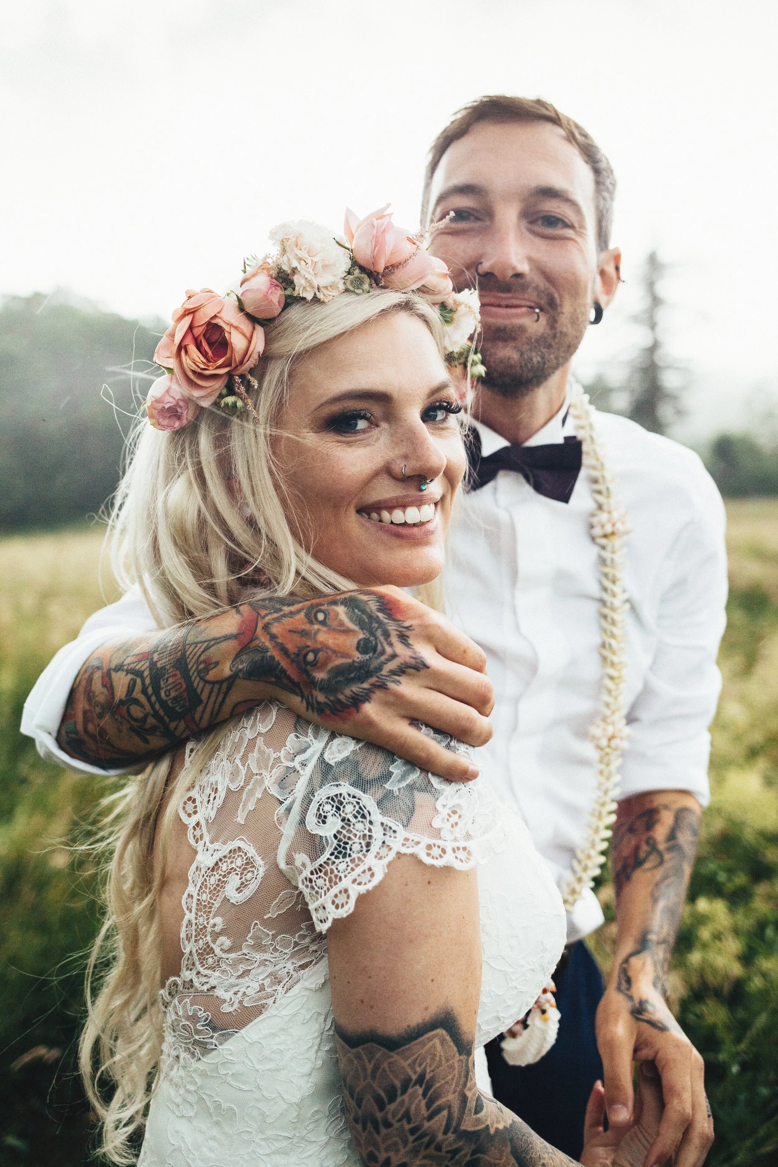 Un couple de mariés en plein air, la femme porte une couronne de fleurs et une robe en dentelle, l'homme porte une chemise blanche avec un collier de fleurs, ils s'étreignent et sourient.
