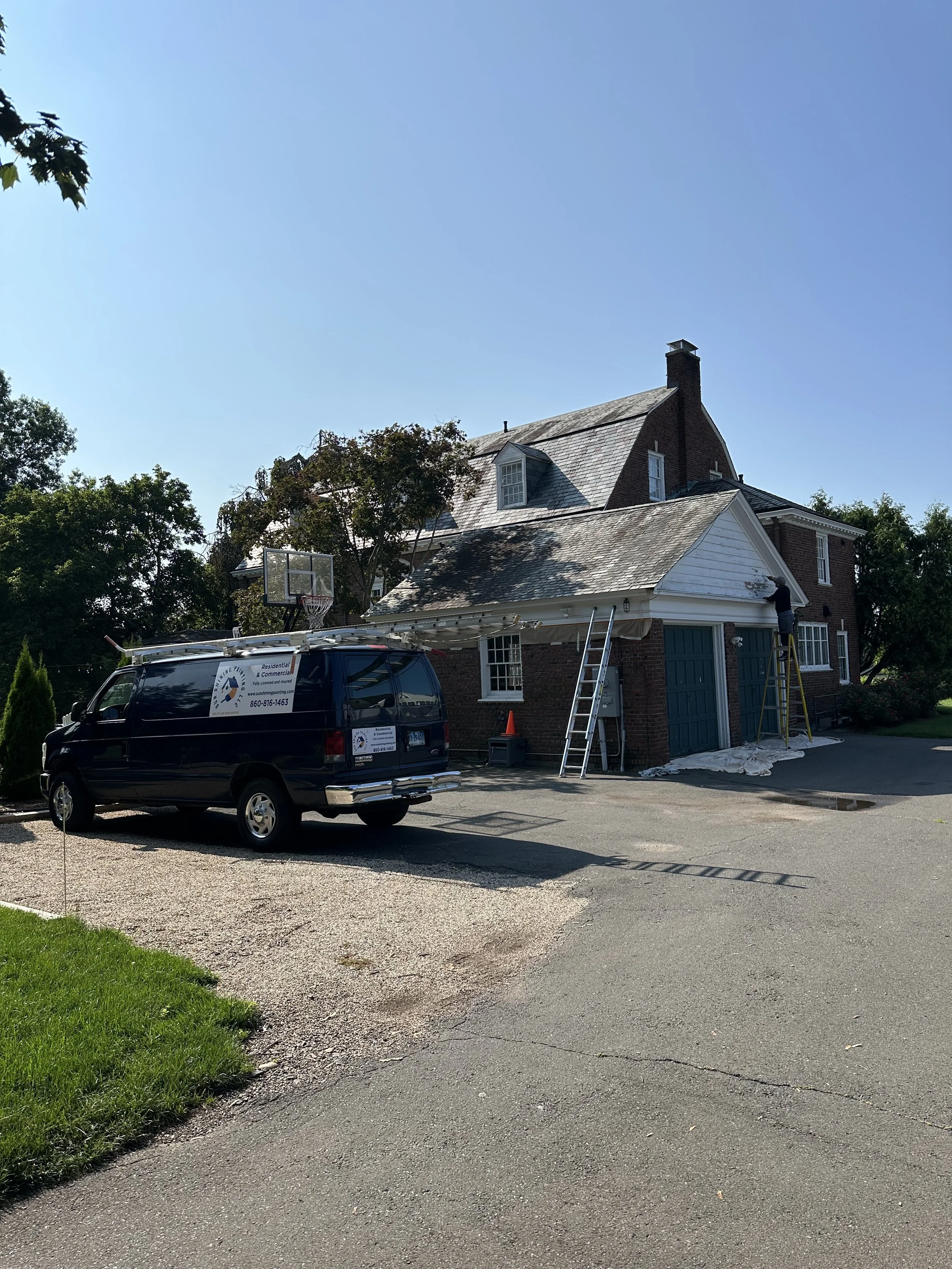 A house undergoing roof repair with workers, ladders, and a van parked in front, in a suburban neighborhood with trees.