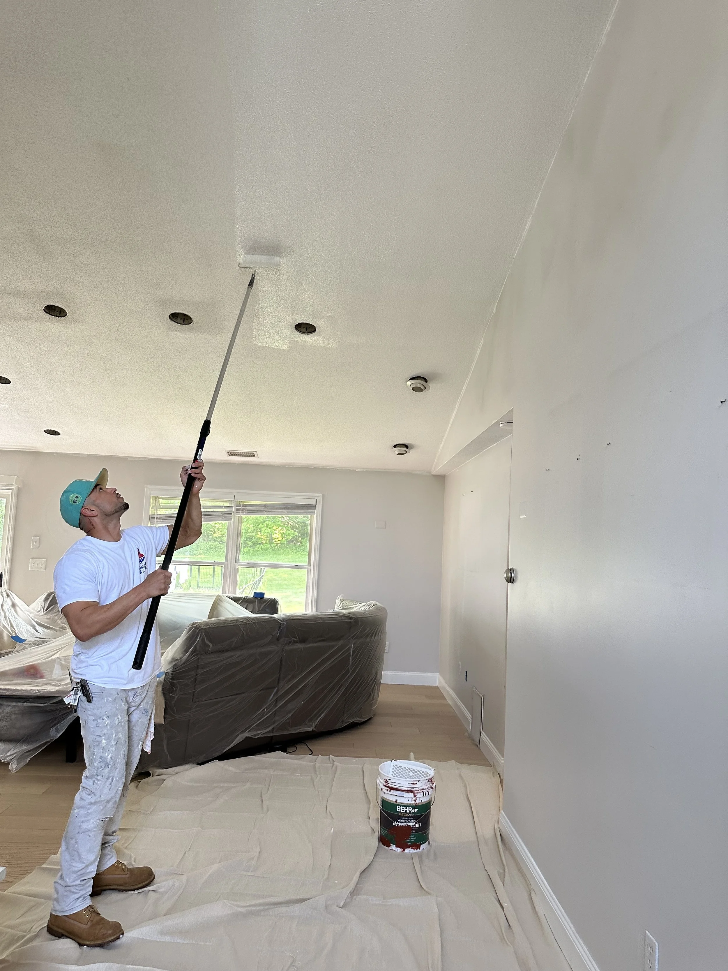A person spray painting a textured white ceiling in a room with large window, beige walls, hardwood floor, and furniture covered with plastic, using a paint sprayer attached to a long pole.
