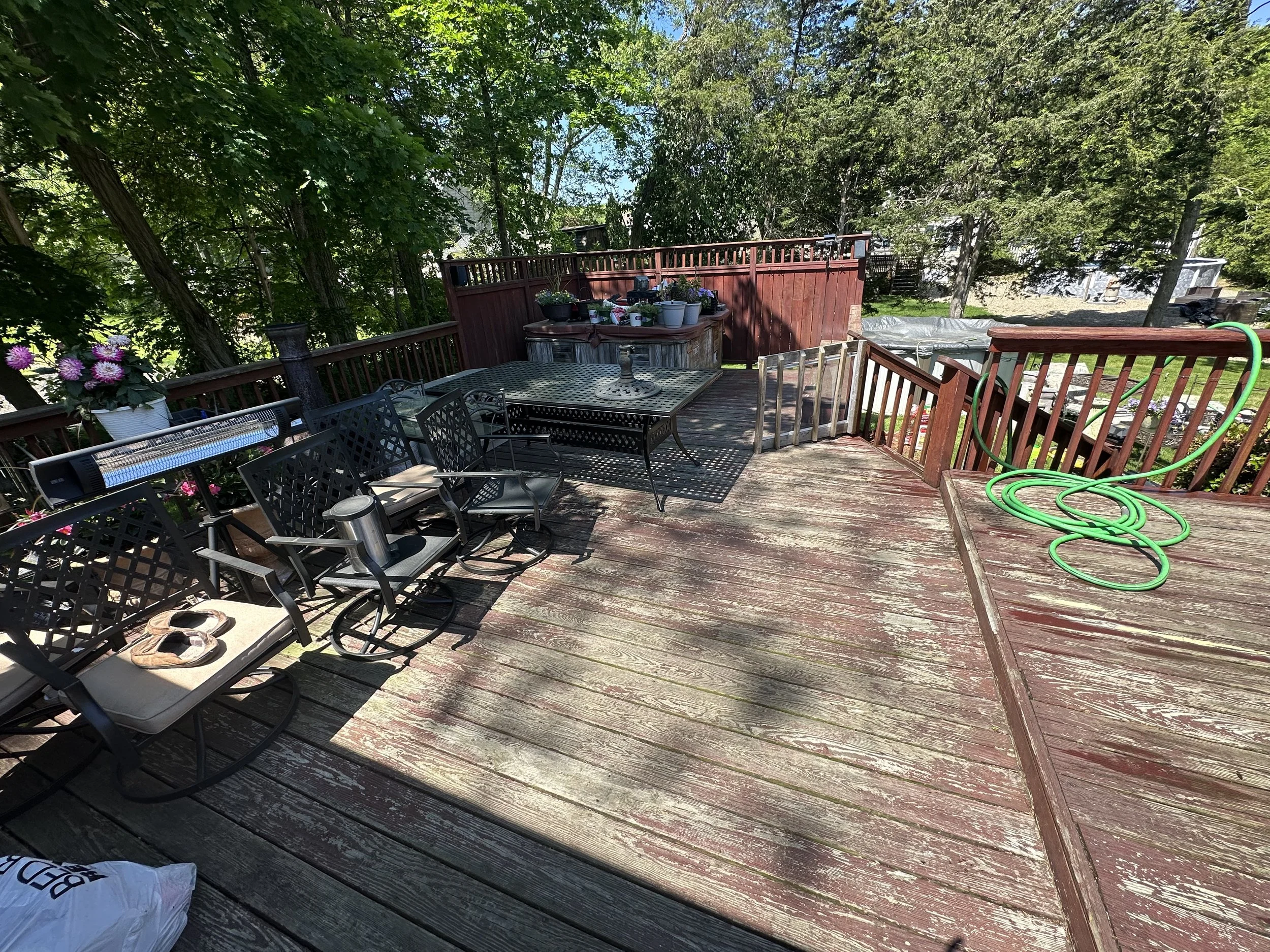 A backyard deck with patio furniture, potted plants, and gardening supplies, surrounded by lush green trees and a wooden fence.