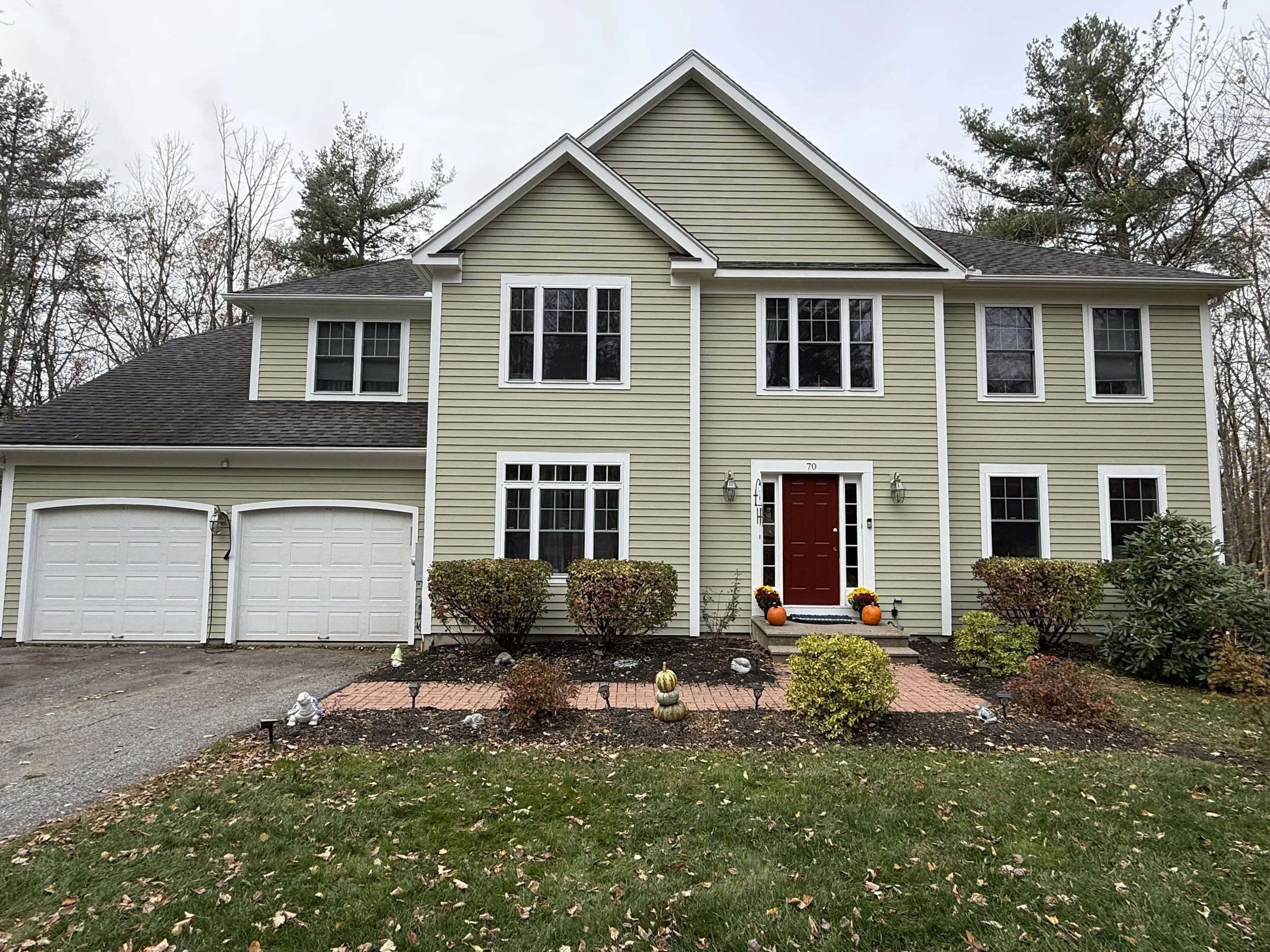 A two-story house with light yellow siding, white trim, and a red front door, decorated with pumpkins and fall plants outside, surrounded by trees.