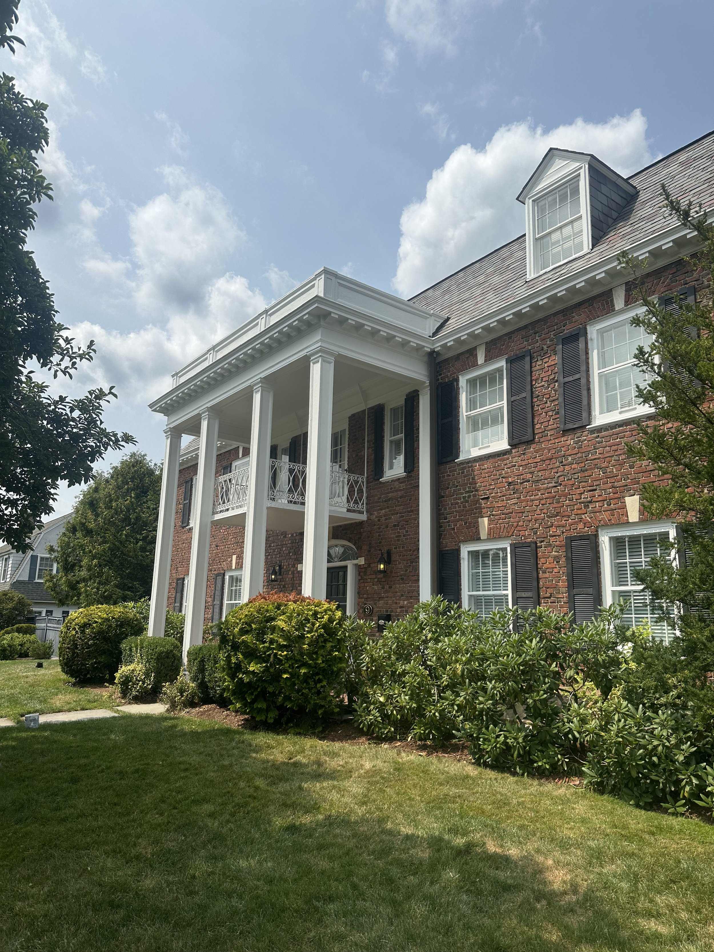 A brick house with white columns supporting a balcony and a dormer window on the roof, surrounded by well-maintained bushes and a grassy lawn under a partly cloudy sky.