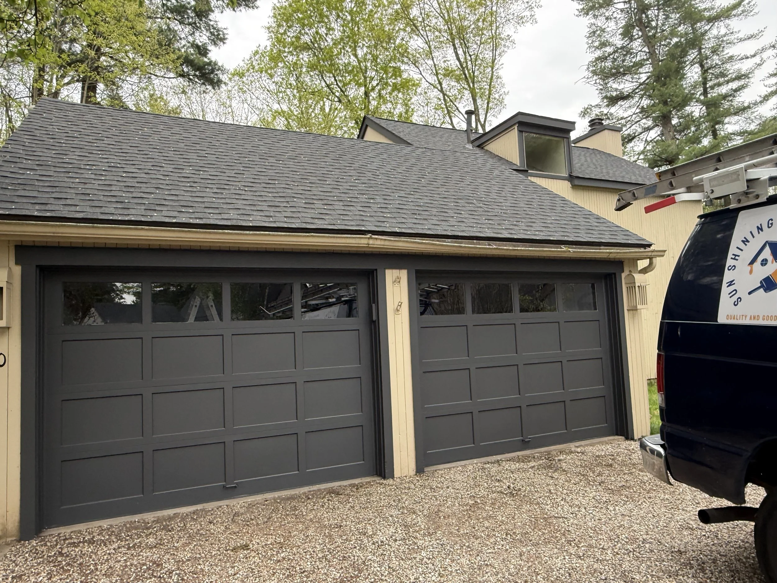 A residential house with a dark gray garage door and a shingled roof. Part of a building with a windowed dormer and chimney visible. Trees with green foliage and a cloudy sky. A white van with a ladder on top is parked beside the garage.