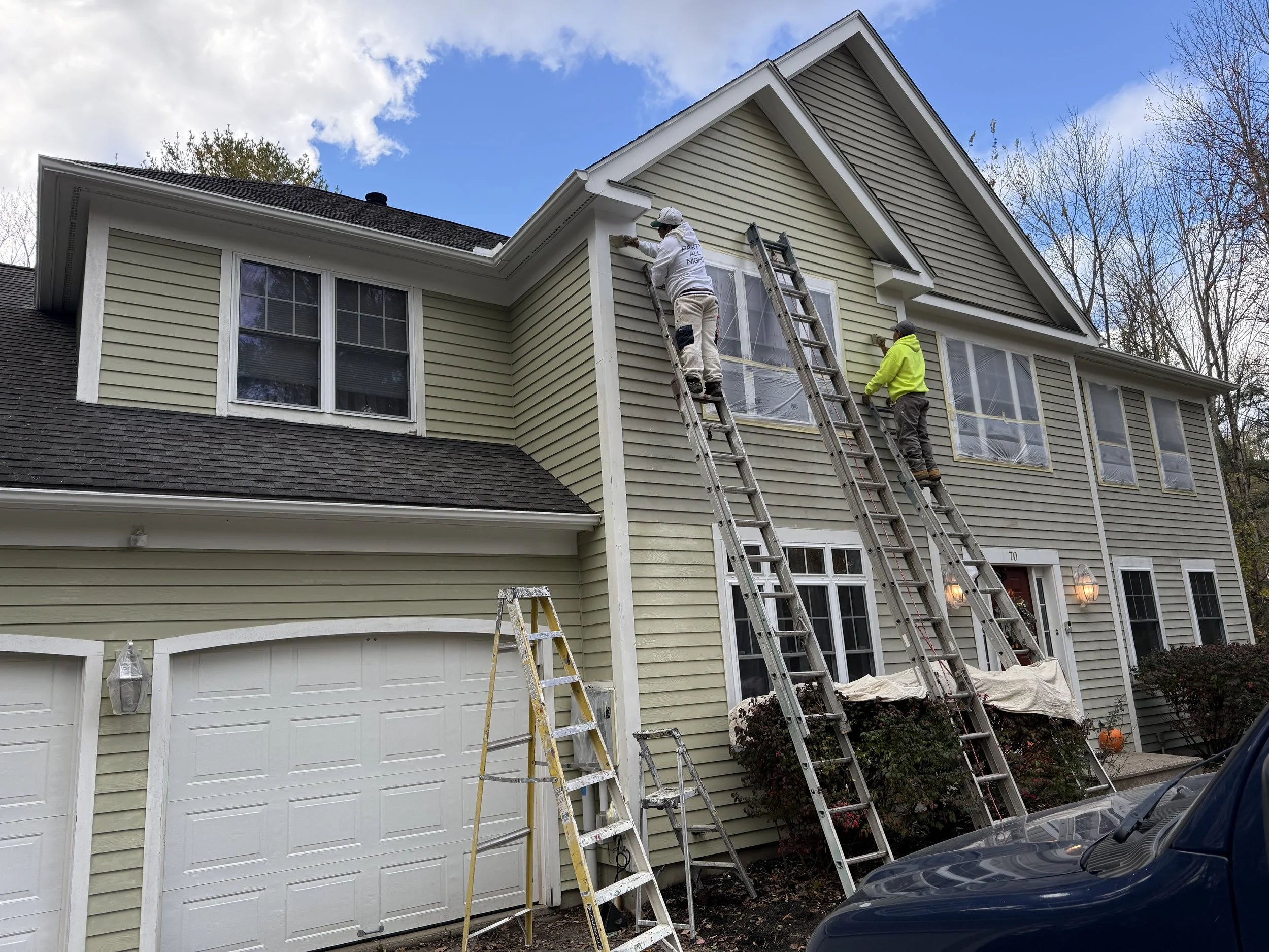 Two workers are painting the exterior of a two-story house, standing on ladders, with one worker reaching near the roof and the other near a window. The house has light yellow siding, multiple windows, and a white garage door beneath. The sky is part