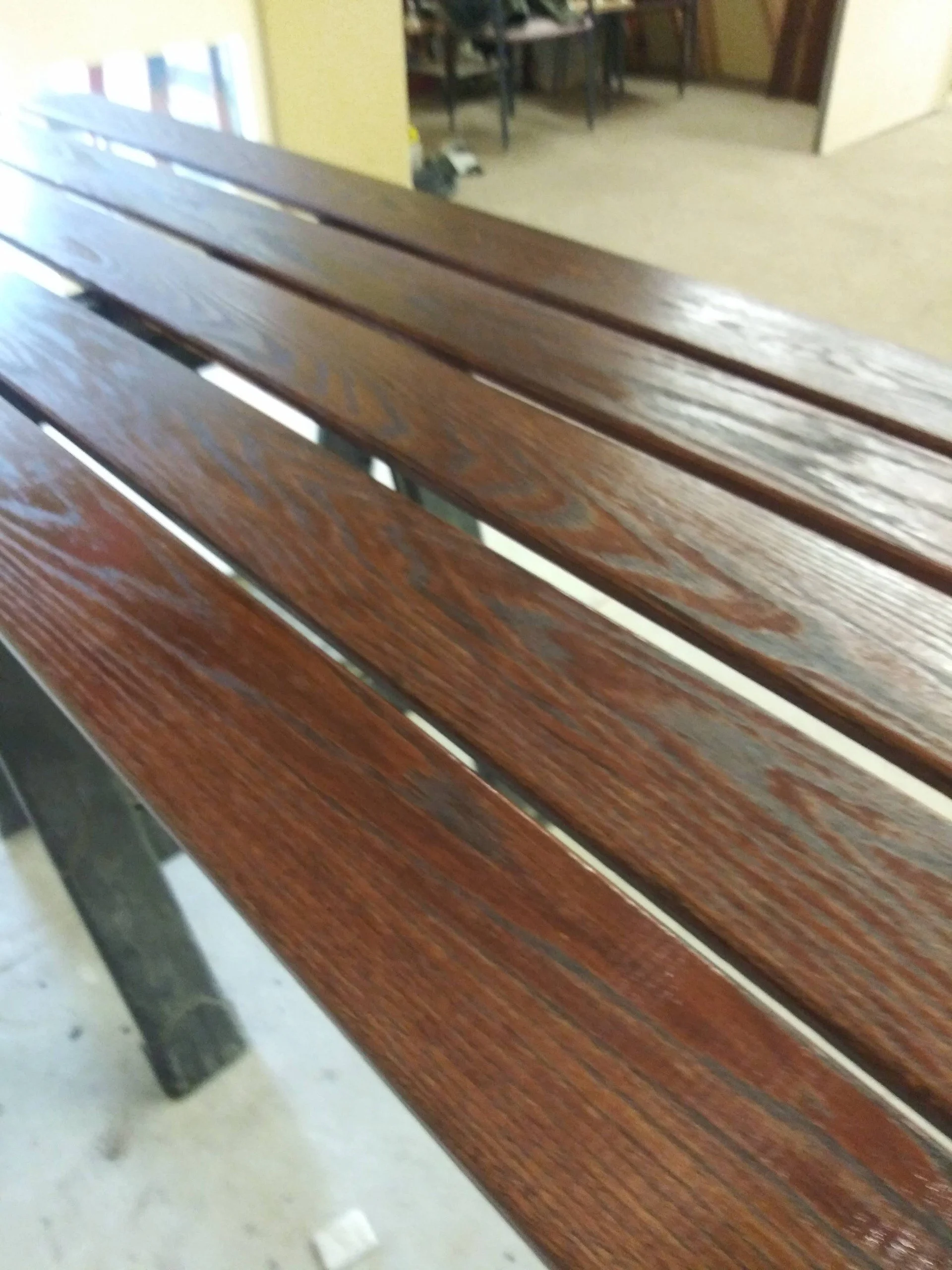 Close-up of a wooden bench showing its brown planks, with a piece of white paper or trash on the floor beneath, in a room with yellow walls and open doorways leading to other rooms.