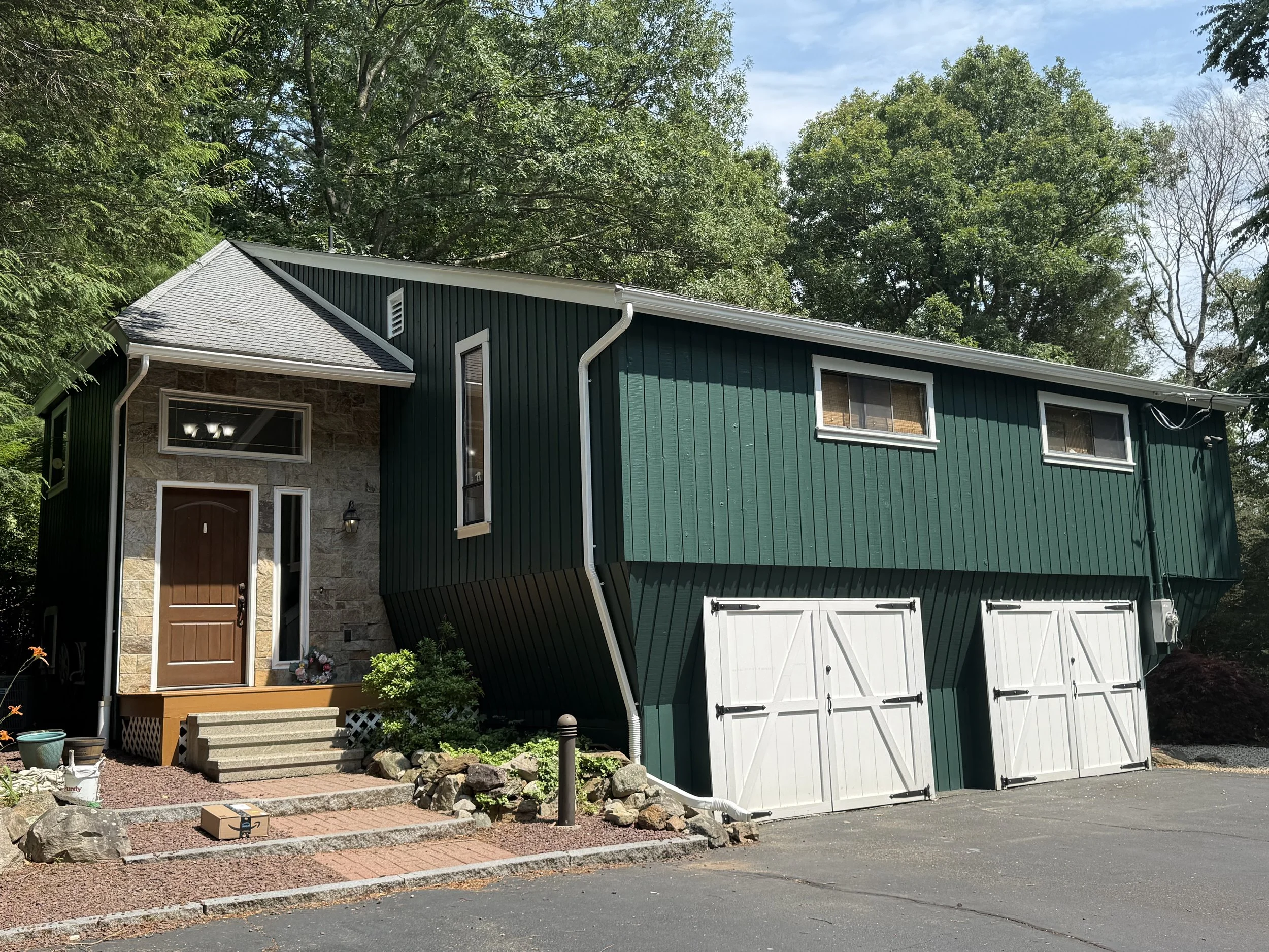A multi-story house with green vertical siding, stone accents, and white garage doors, surrounded by trees and a small front garden.
