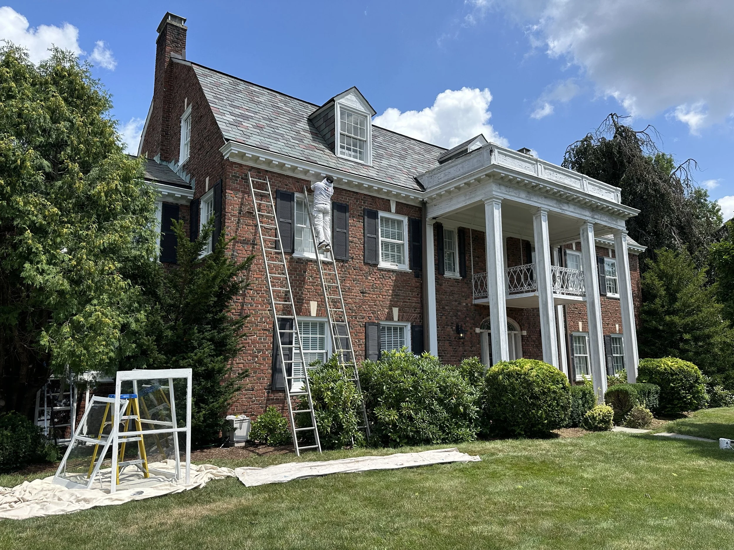 Person on a ladder working on the roof of a brick house with white trim, black shutters, and a porch, surrounded by green trees and a blue sky.
