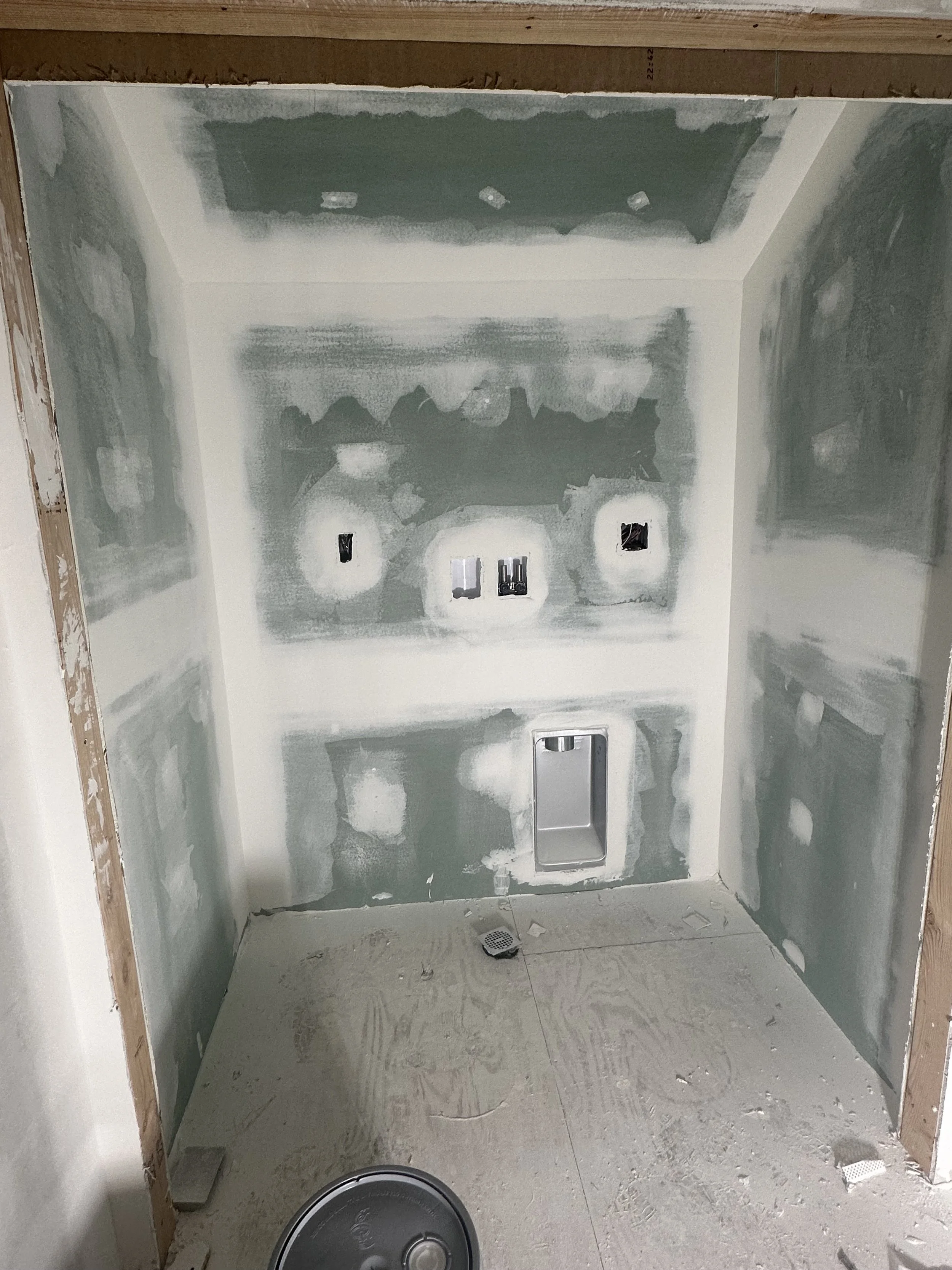 Unfinished shower with drywall and electrical outlets, construction debris on the floor, and a drain in the center.