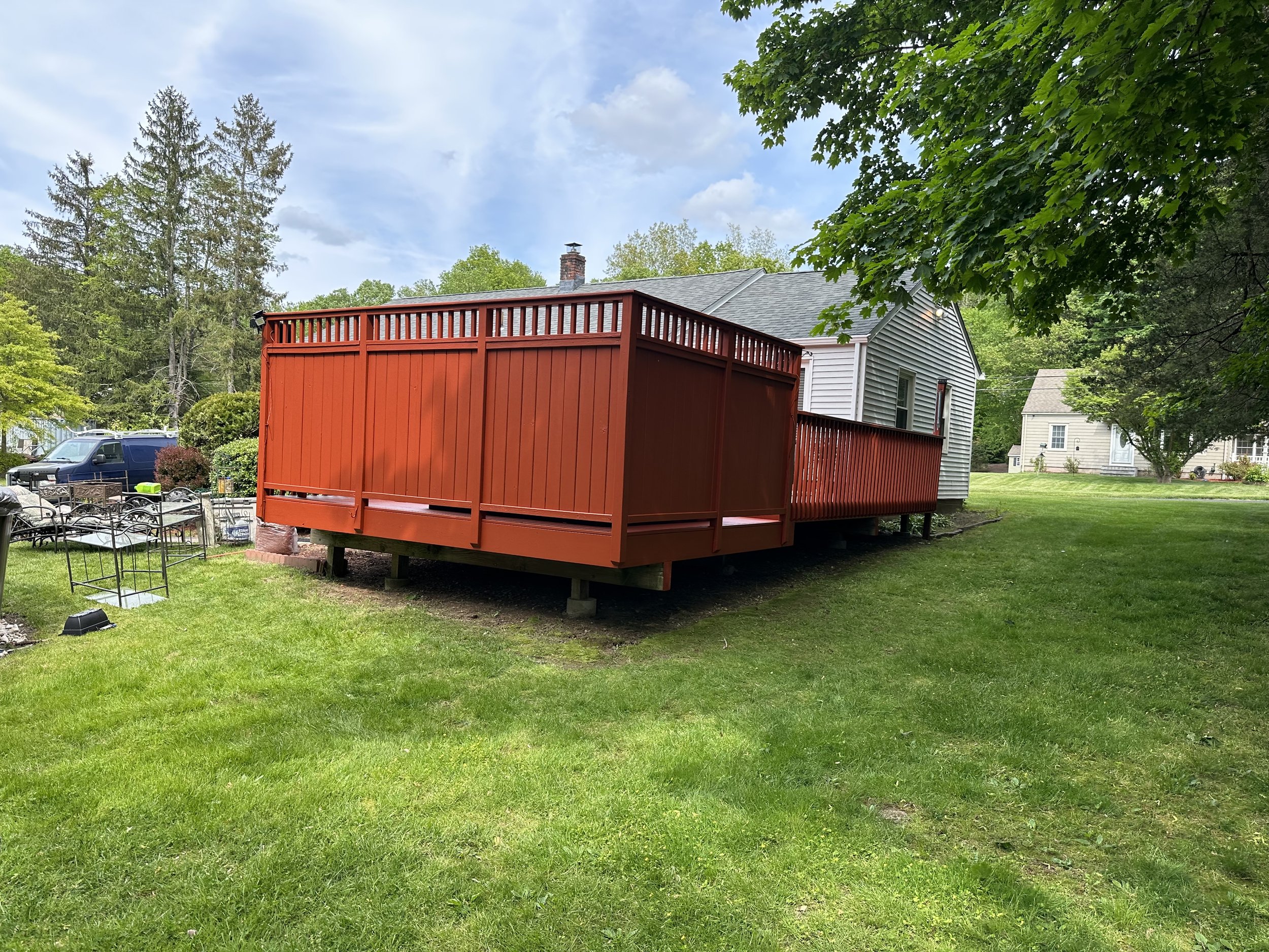 A backyard with a red deck attached to a gray house, green grass, trees, and a partly cloudy sky.