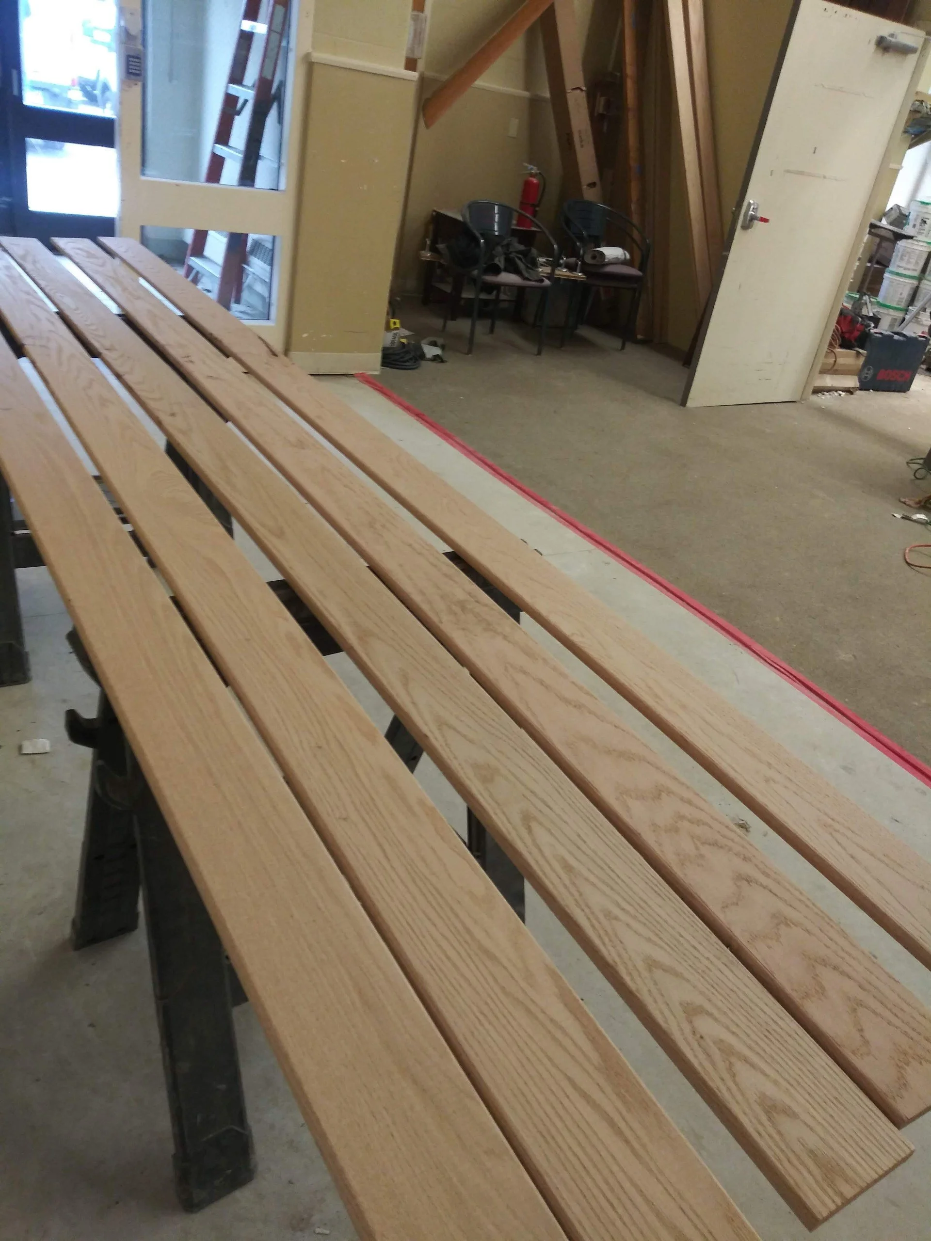 A wooden slat bench in a workshop or store, with a ladder and chairs in the background.