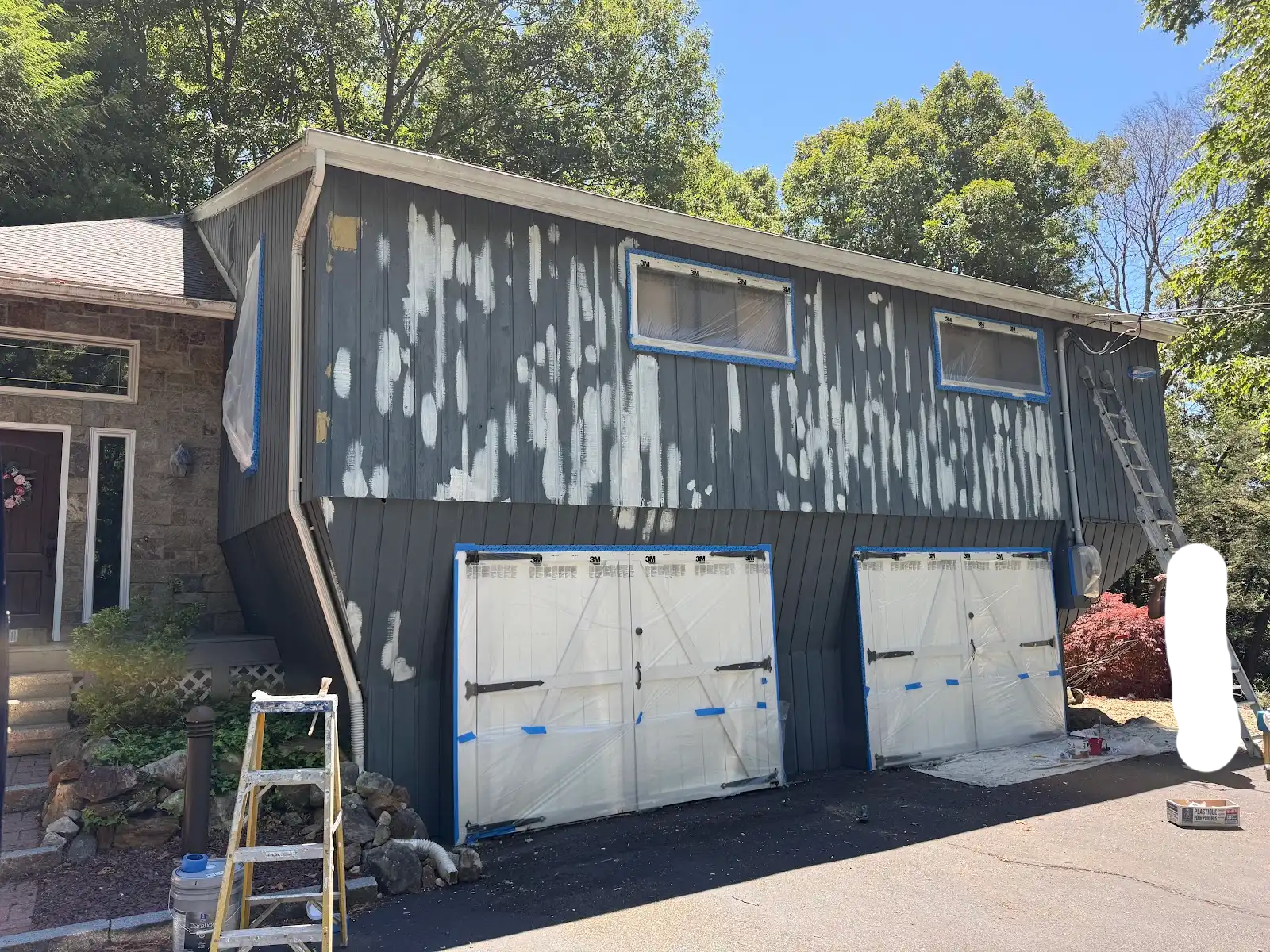 A house undergoing exterior renovation with siding being painted or replaced. The house has multiple windows covered with protective plastic. A ladder leans against the house, and painting or work supplies are nearby, with partial trees and a blue sk