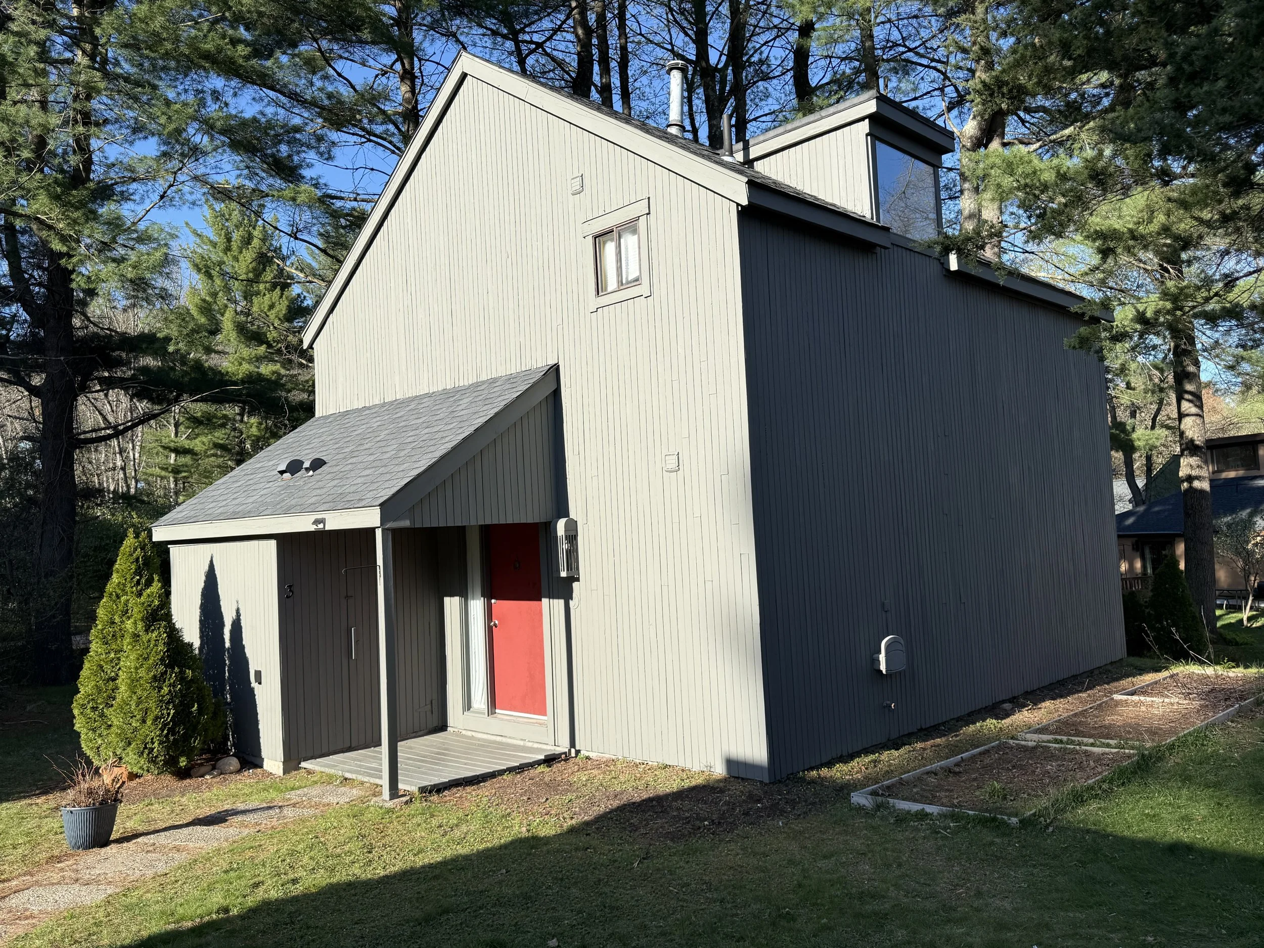 A modern, multi-story house with vertical siding, a red front door, and a small front porch, surrounded by trees and a well-kept yard.