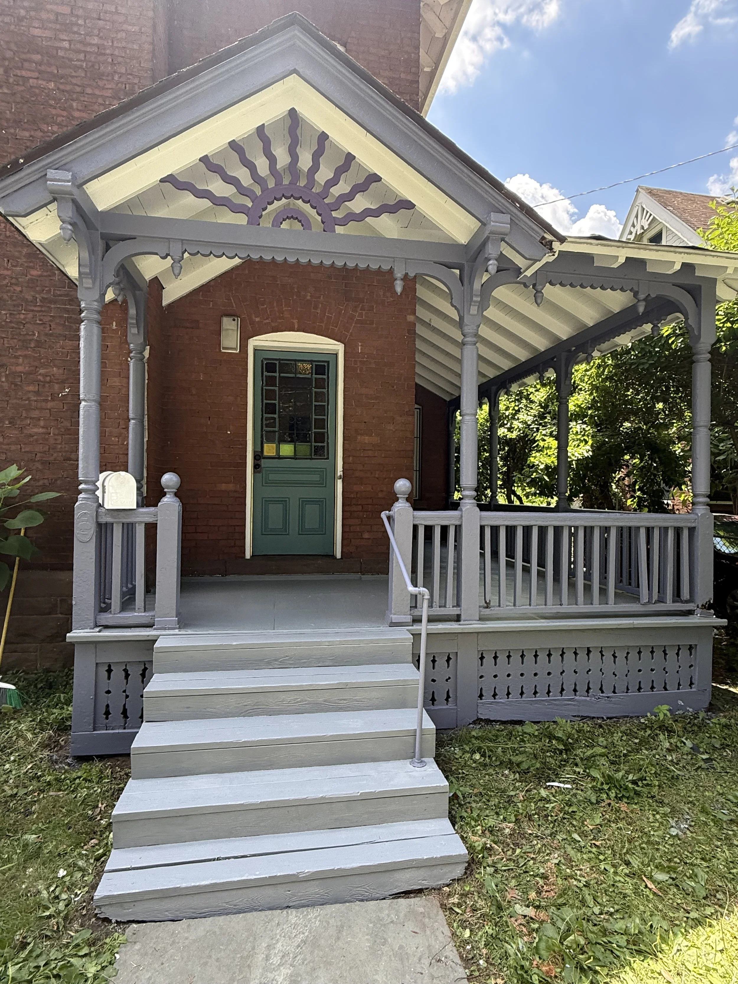 Front porch with steps, railing, and decorative trim, attached to a brick house with a green door and small window.