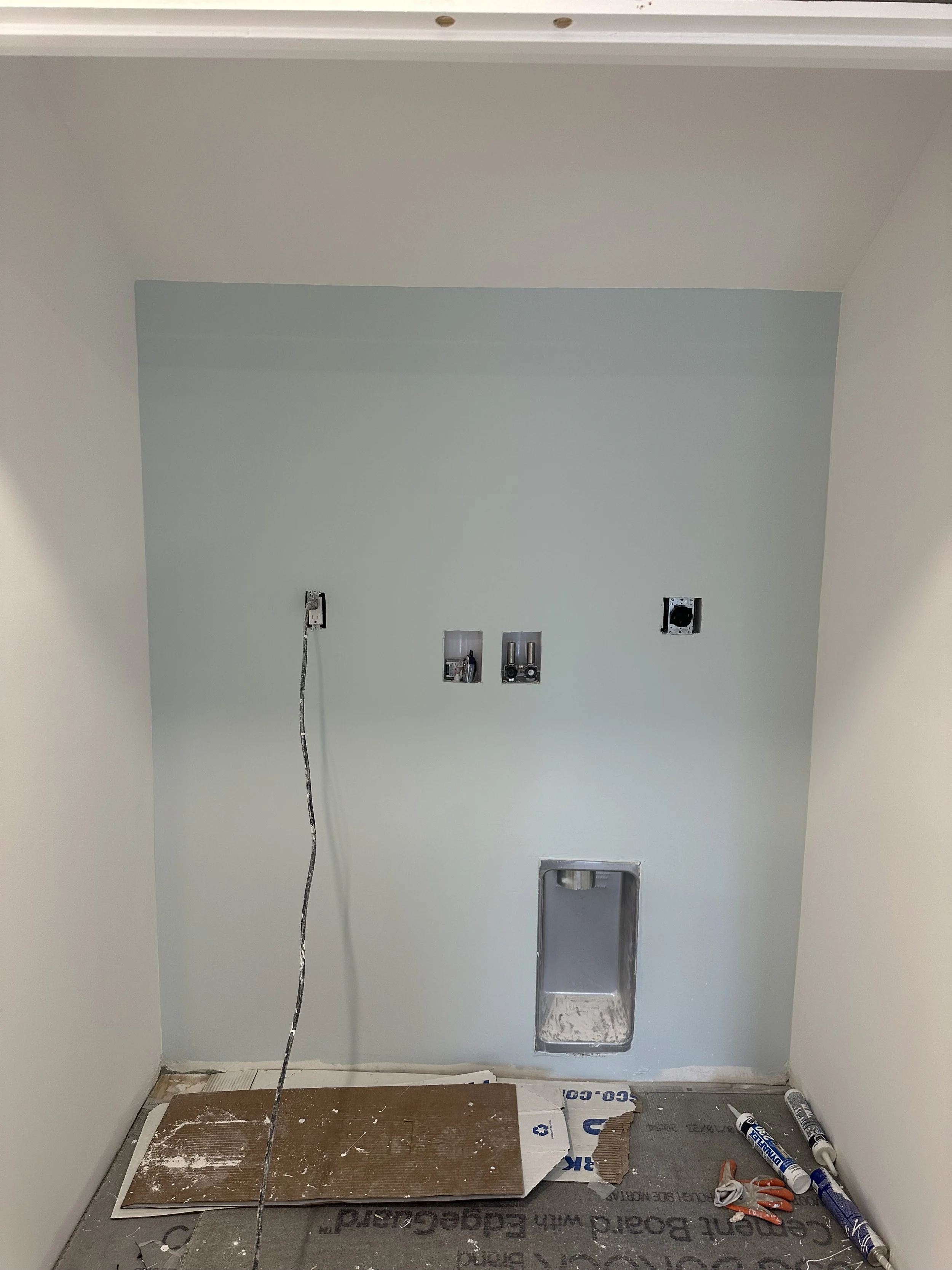 A room under renovation showing a light-colored wall with electrical outlets and wiring, a small drain on the floor, construction materials, tools, and protective covering on the floor.