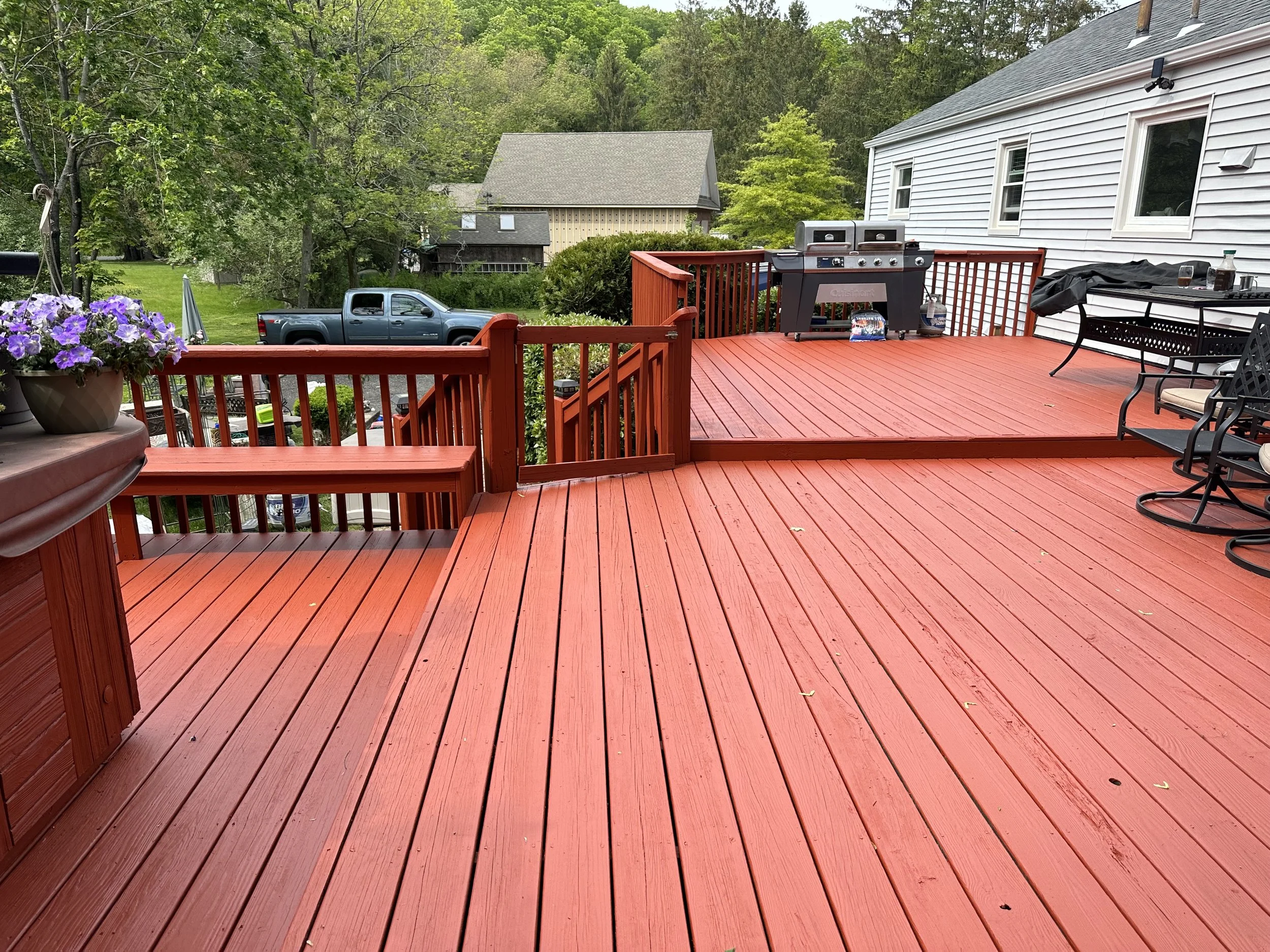 A red wooden deck with a barbecue grill, table, chairs, and a flower pot. A gray truck is visible in the background, along with green trees and neighboring houses.