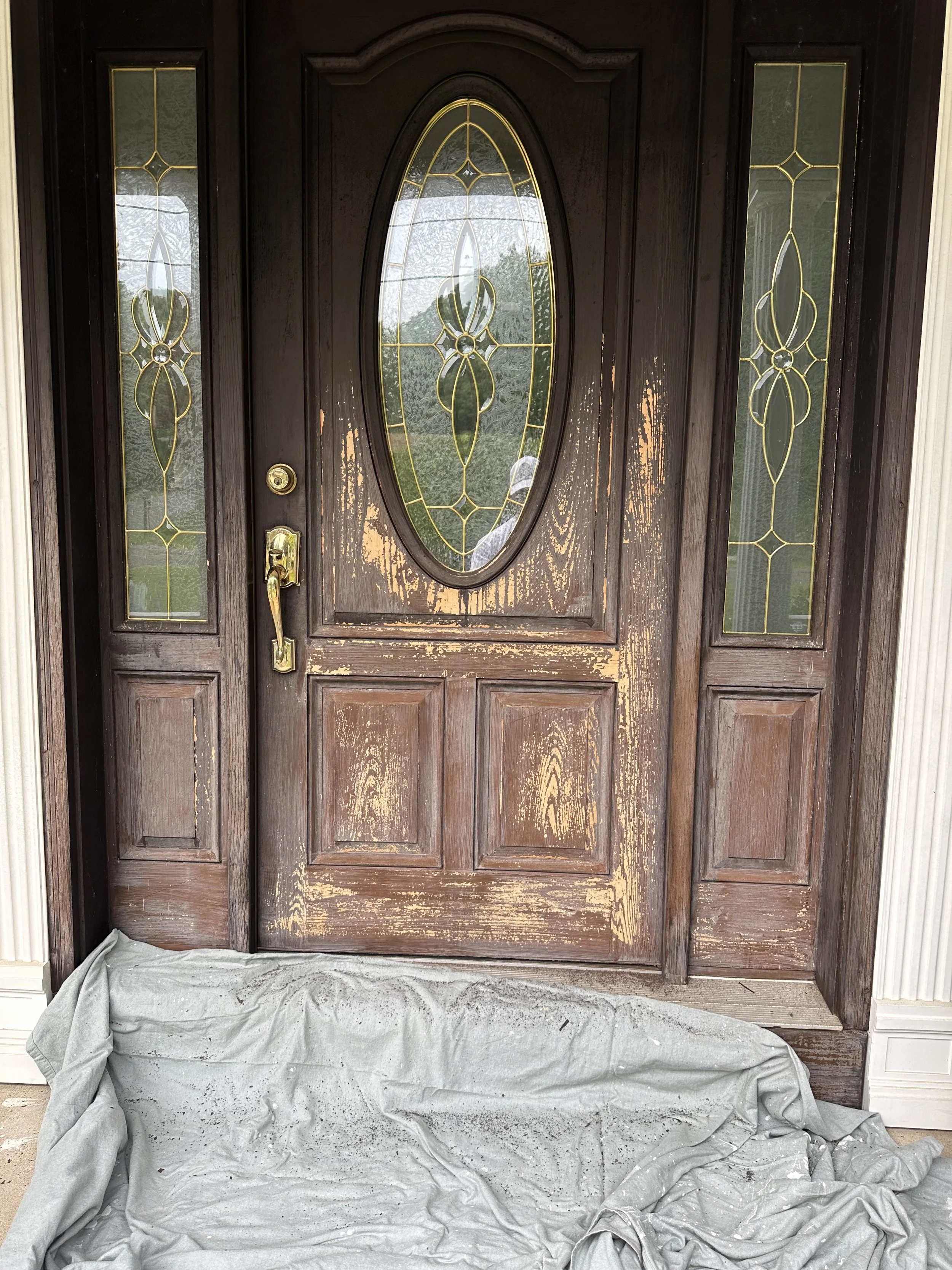 A wooden front door with decorative glass panels on each side and an oval glass window in the center. The door has a brass handle and lock, and the lower part shows signs of being recently sanded or refinished. A sheet covers the threshold and part o