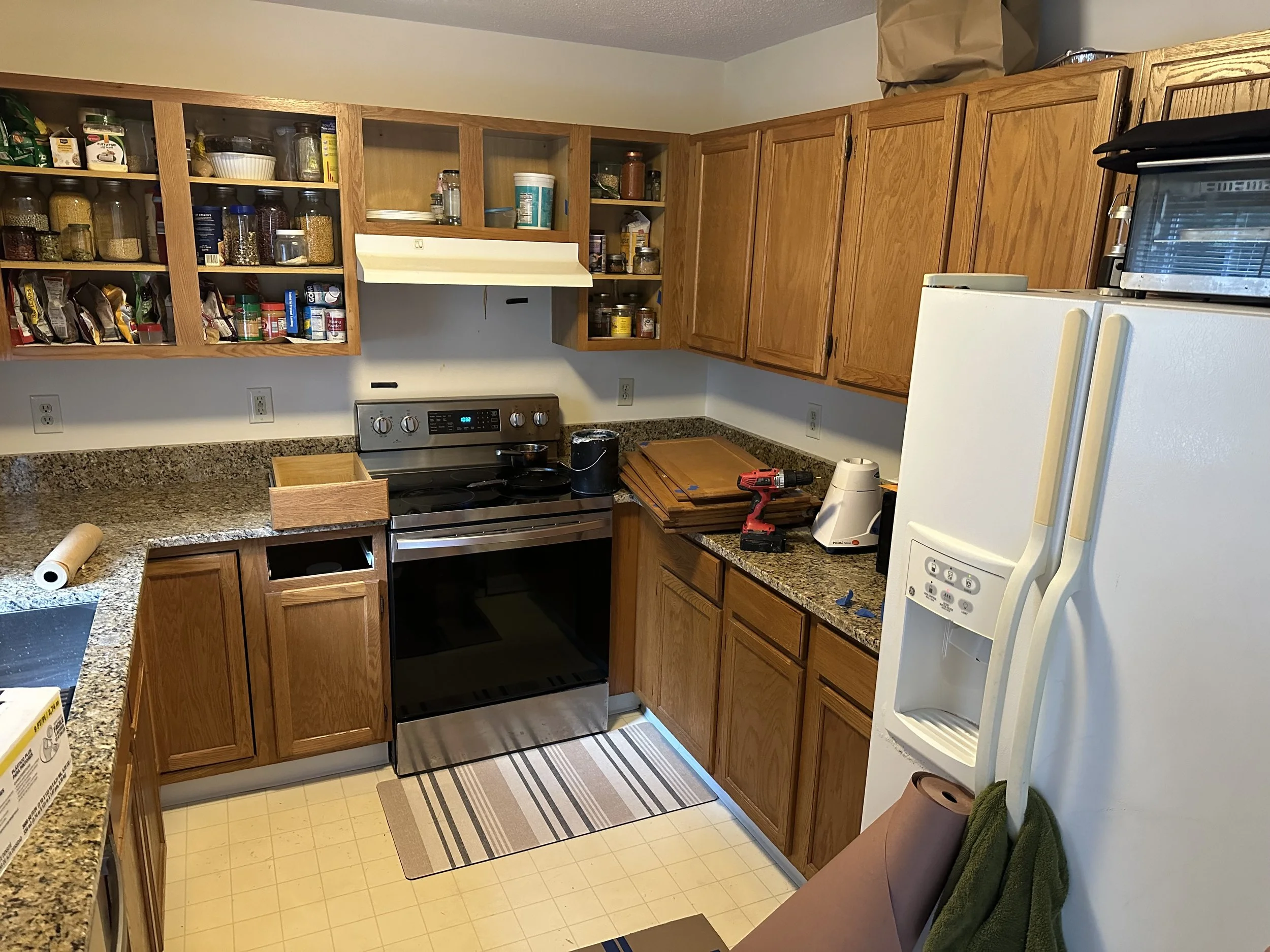 A kitchen with wooden cabinets, open shelves filled with pantry staples, a stove, a refrigerator, and various kitchen utensils on the countertop.