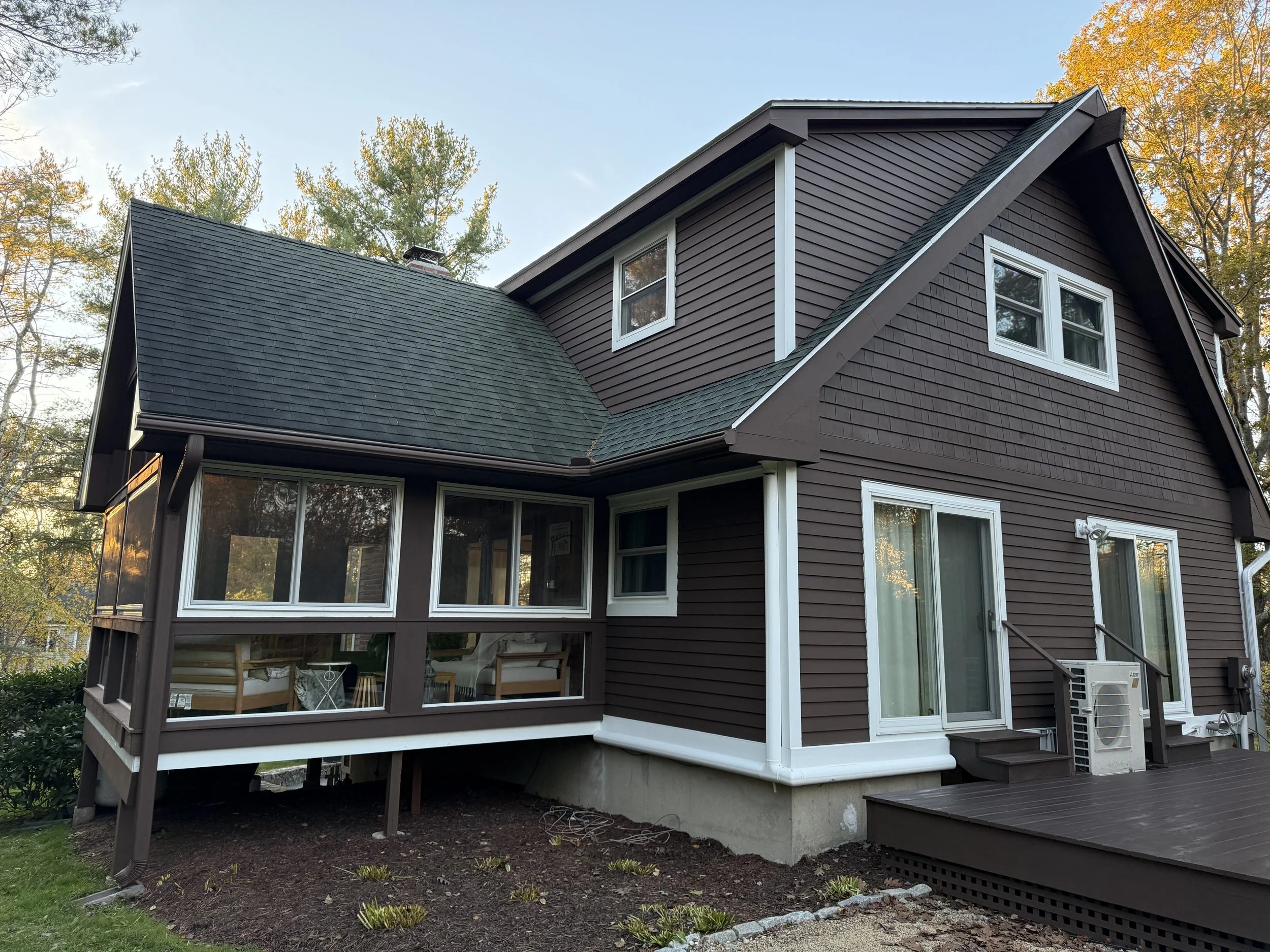 The image shows the upper part of a modern two-story house with brown siding and white trim, surrounded by trees with fall foliage. The house features multiple windows, a sloped roof, and a screened-in porch area at the bottom.