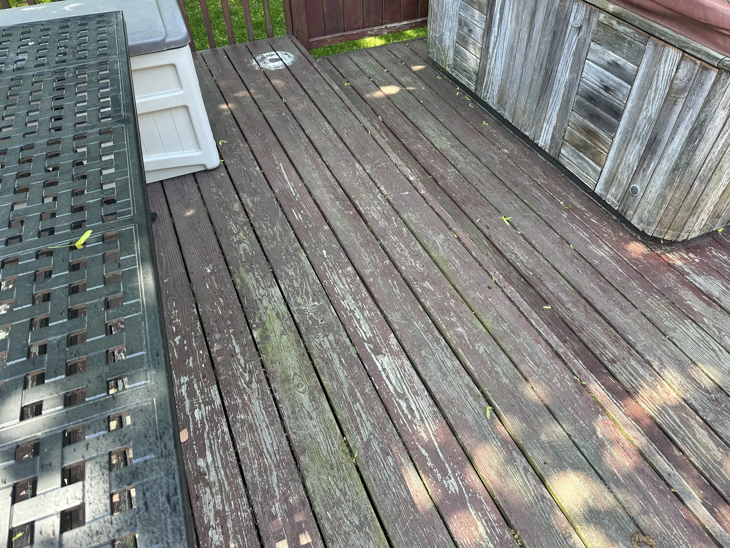A wooden deck with weathered, reddish brown planks, with a metal table on the left side and a white plastic step stool nearby. There are some small green leaves scattered on the deck, and a part of a wooden railing and wall are visible.