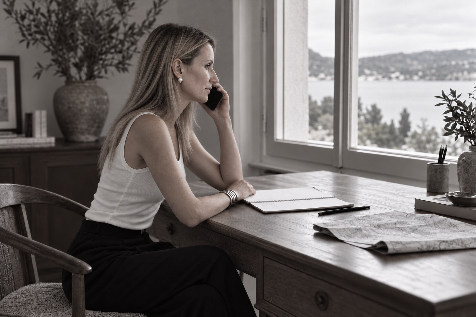 Femme assise à un bureau en train de parler au téléphone, vue latérale, à côté d'une grande fenêtre avec vue sur un paysage en arrière-plan, dedans un décor minimaliste avec des plantes et des livres.