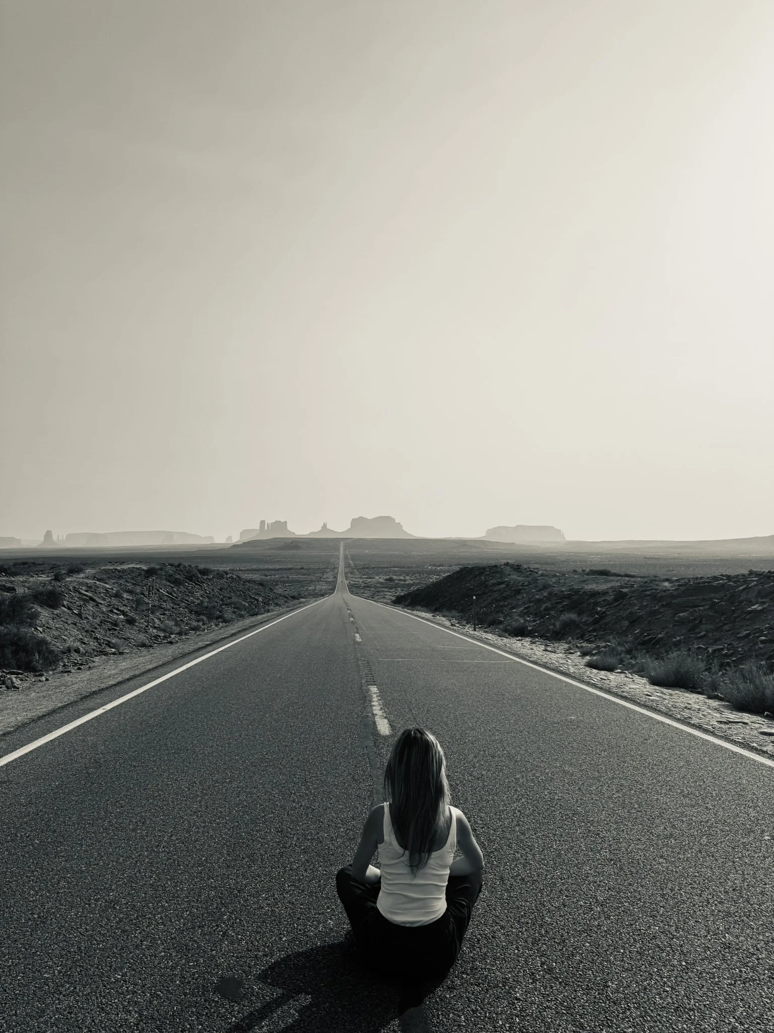 Une personne assise au centre d'une route déserte qui s'étend vers l'horizon avec des formations rocheuses en arrière-plan, sous un ciel clair.
