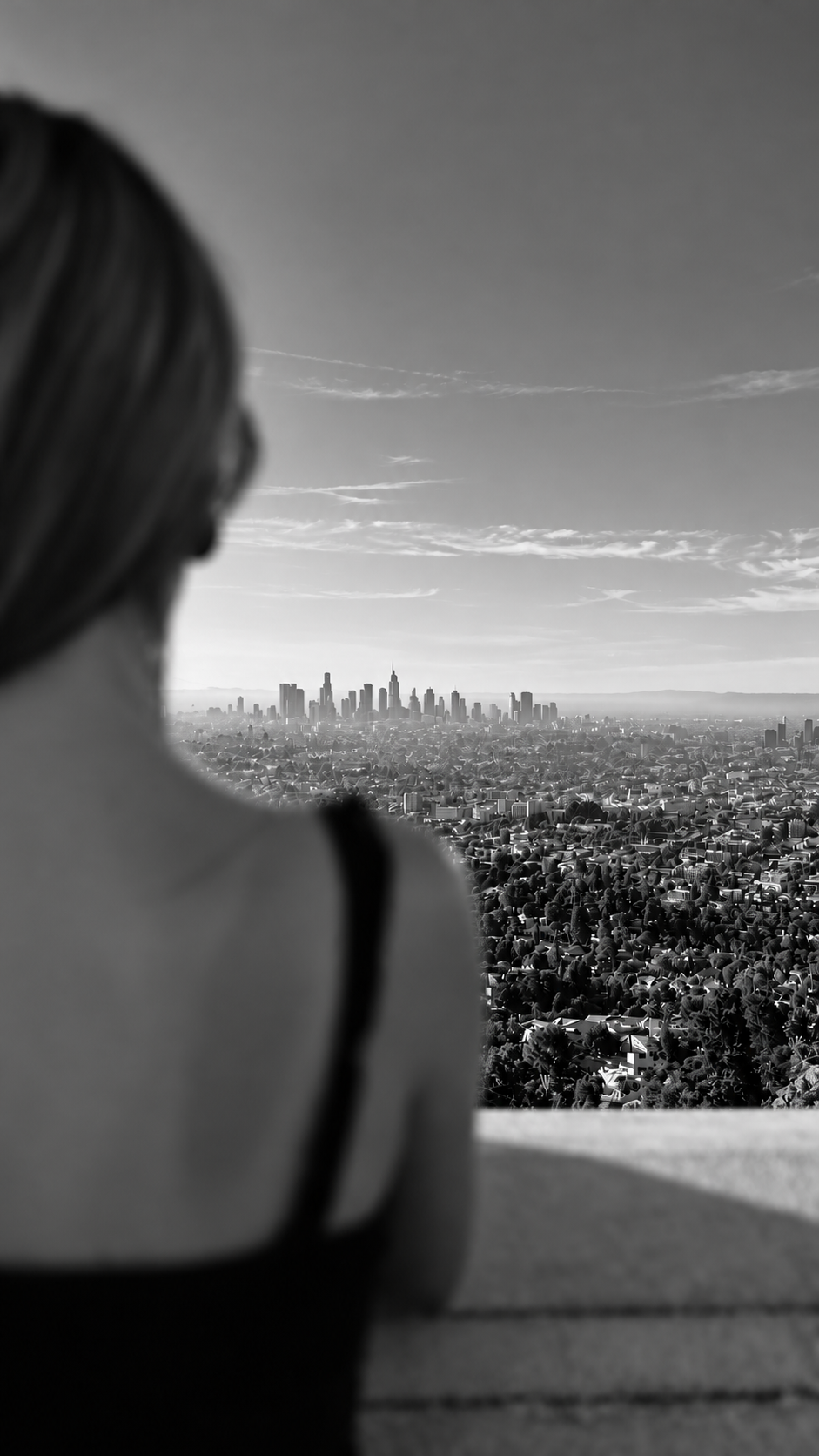 Une femme regarde un skyline urbain à distance, en noir et blanc, depuis un point élevé ou une terrasse.