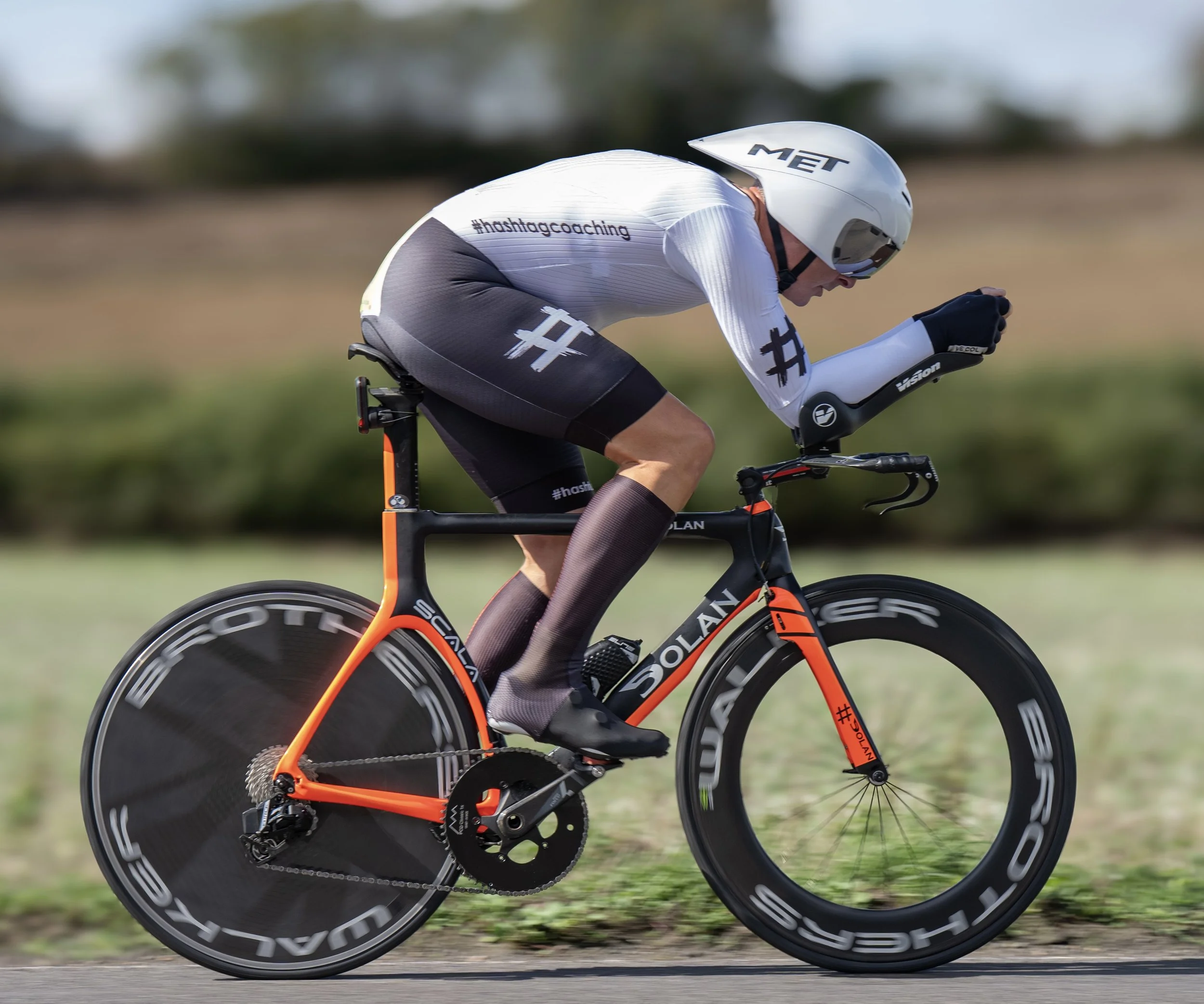 A cyclist riding a black and orange bicycle on a paved path, wearing a white helmet and black sunglasses, with a blurred outdoor background.